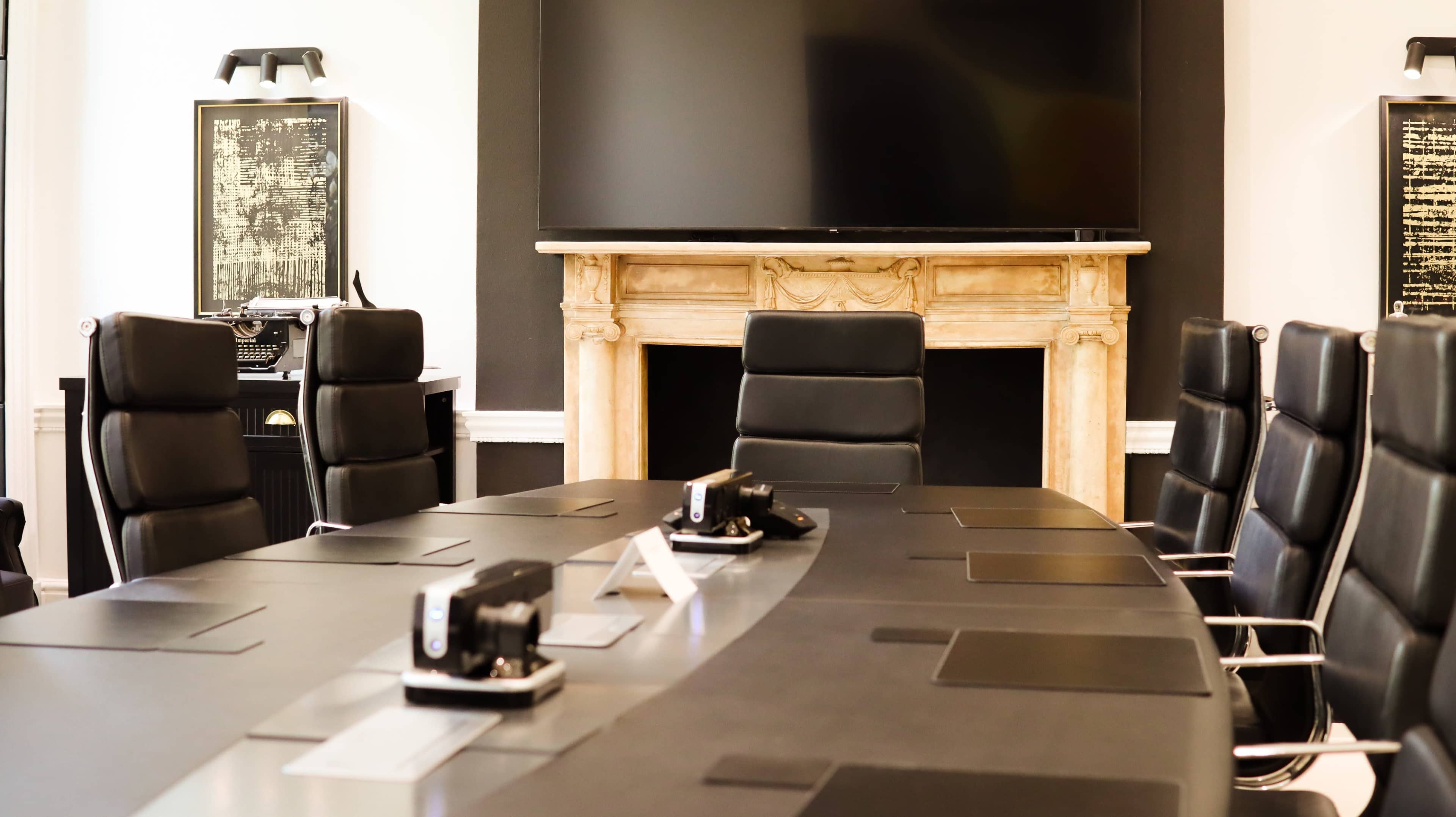A modern conference room features a long, black table surrounded by leather chairs, with a large television mounted on the wall and a decorative fireplace in the background.