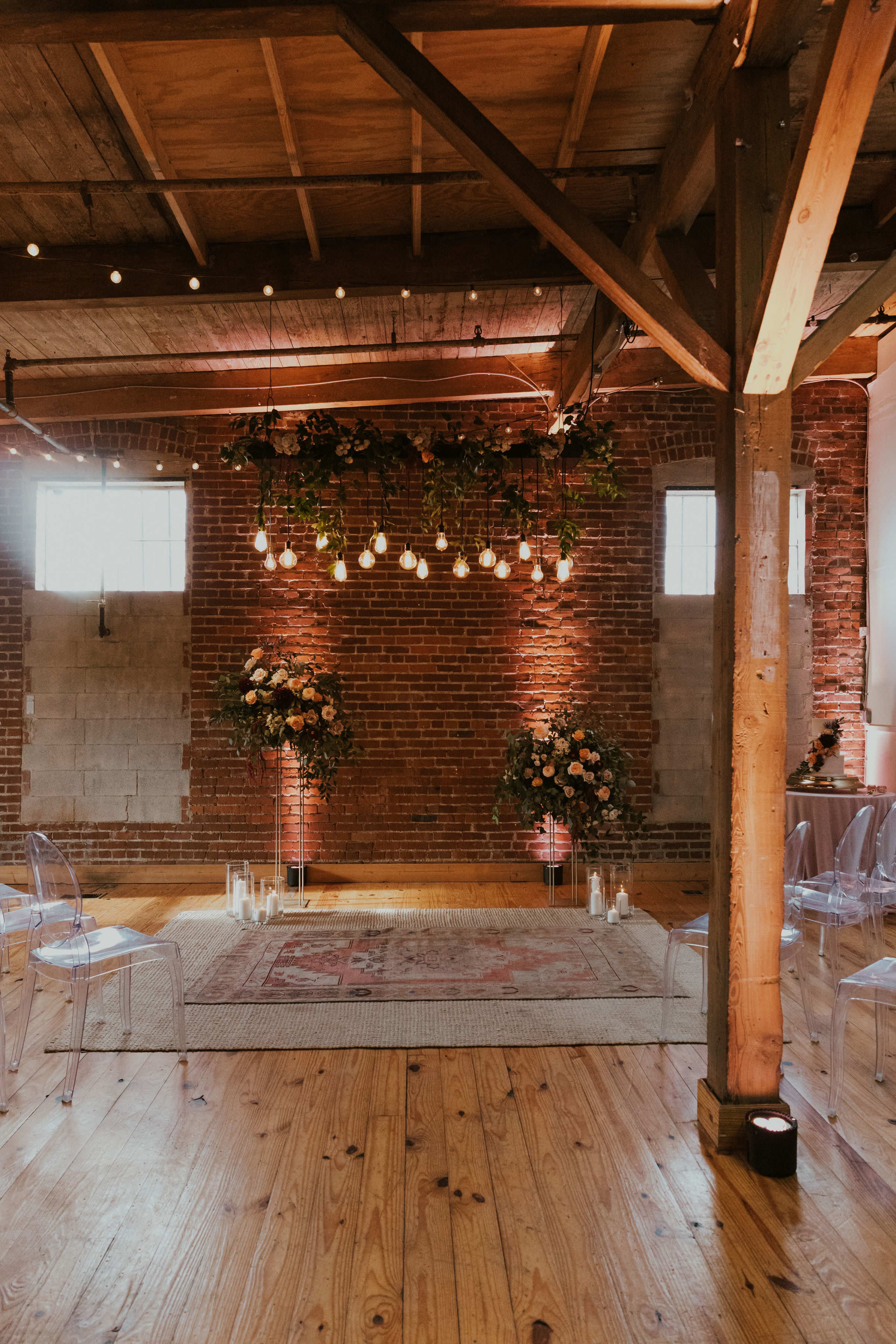 A spacious interior with wooden floors, exposed brick walls, and overhead lights, featuring floral arrangements and clear chairs arranged around a central area.