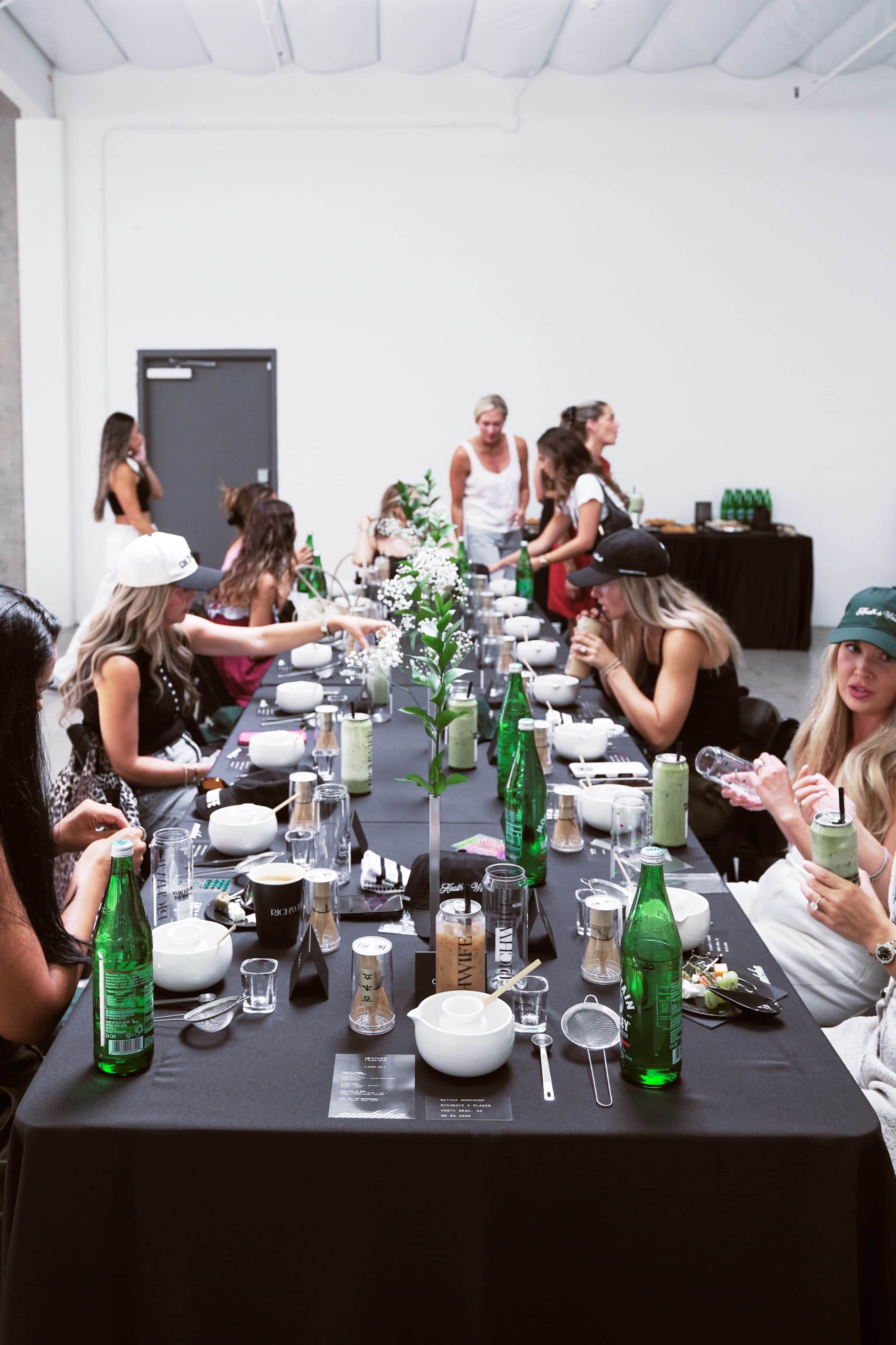 A long table is set for a dining event with multiple guests seated, green bottles on the table, and floral centerpieces.