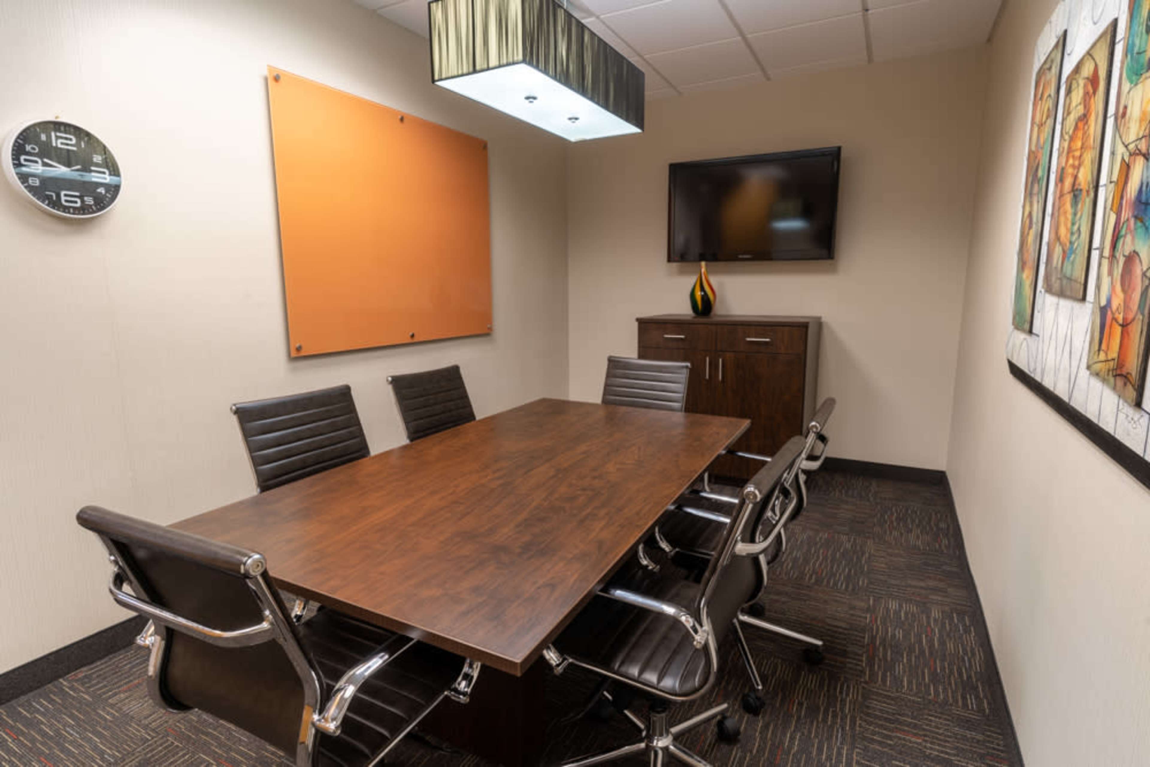 A conference room features a long wooden table surrounded by black leather chairs, a mounted television, and wall art.