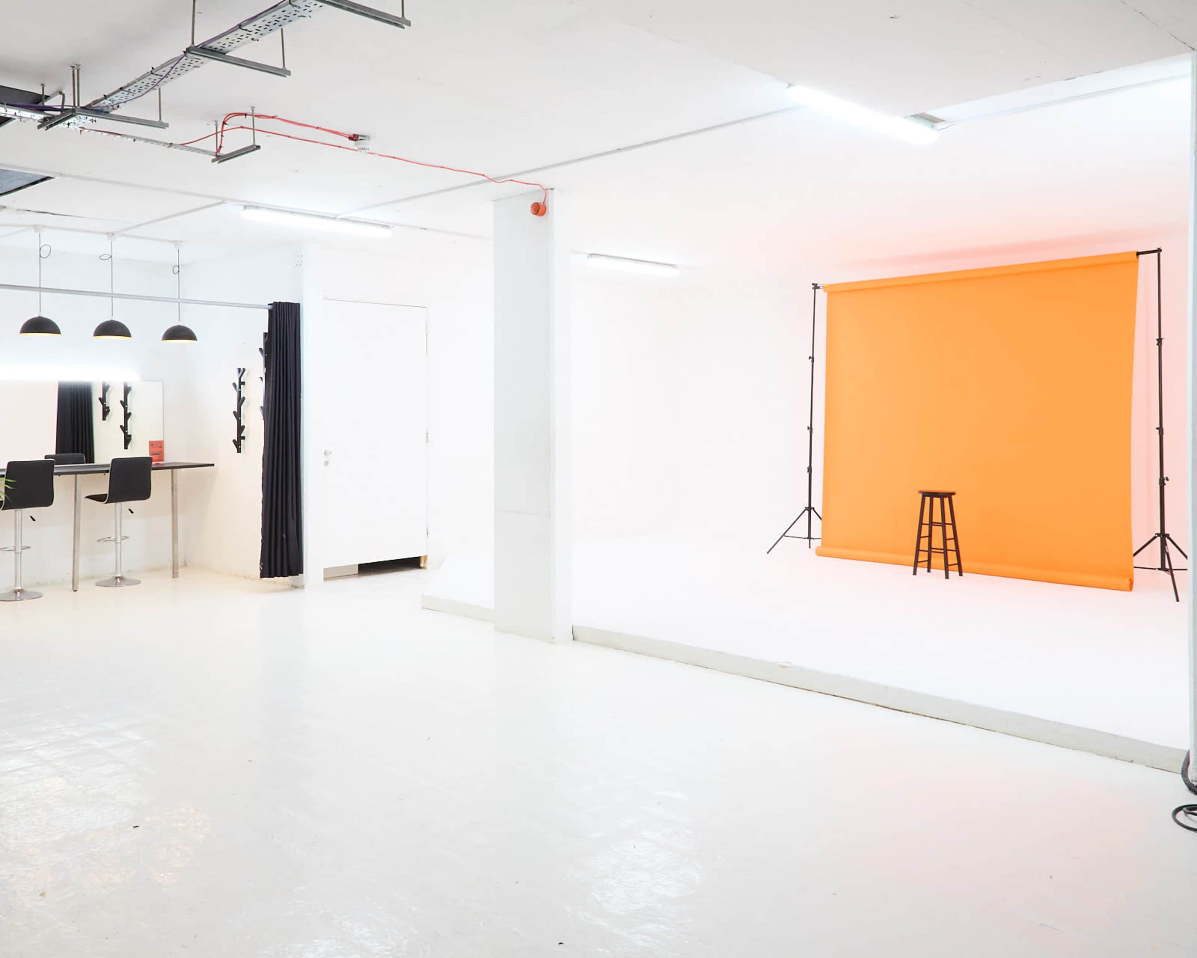 A photography studio featuring an orange backdrop, a black stool, a makeup area with bar stools, and white walls and flooring.