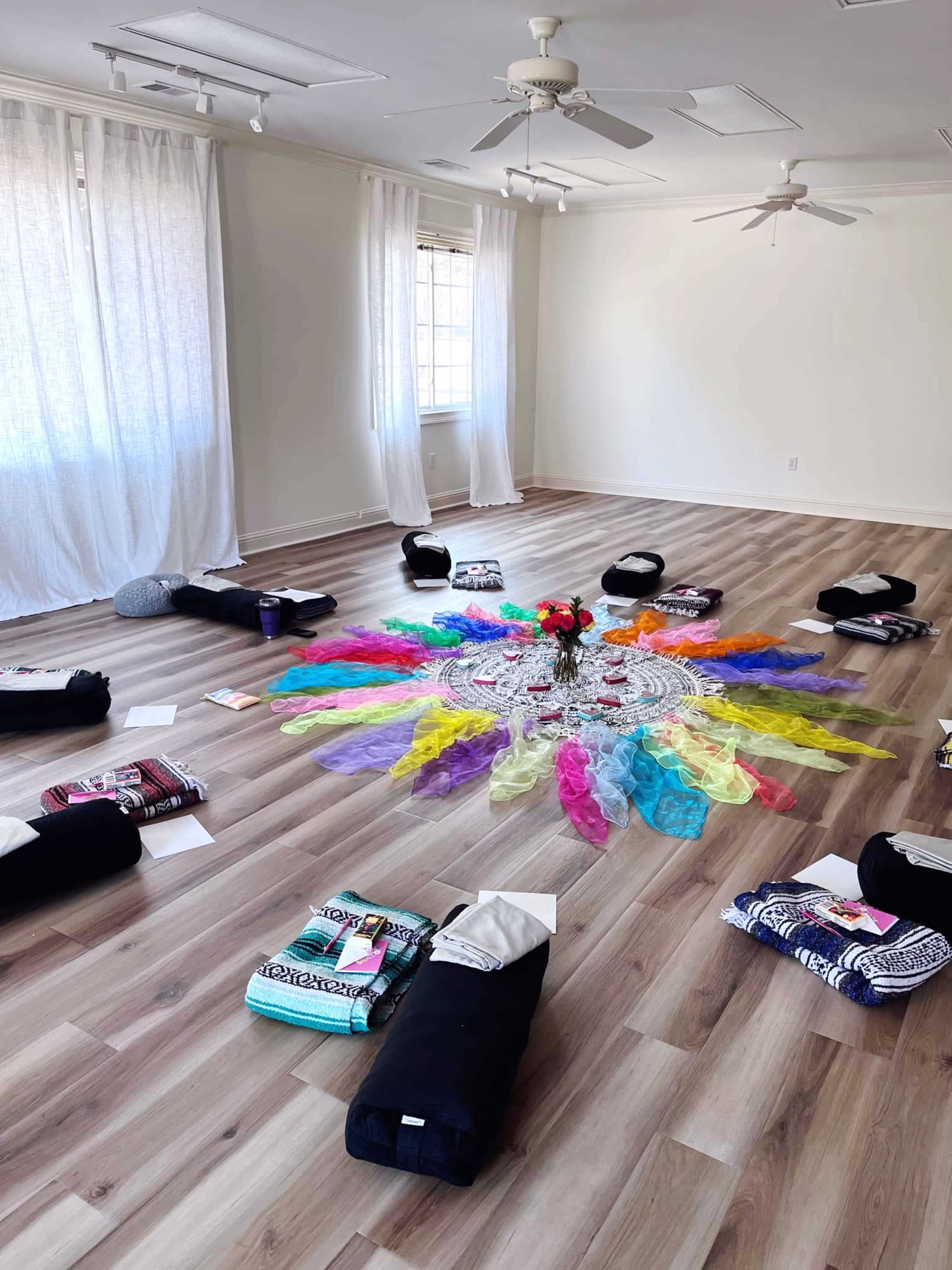 A spacious yoga studio is arranged with yoga mats and props in a circular pattern around a colorful mandala made of fabric and flowers at the center.