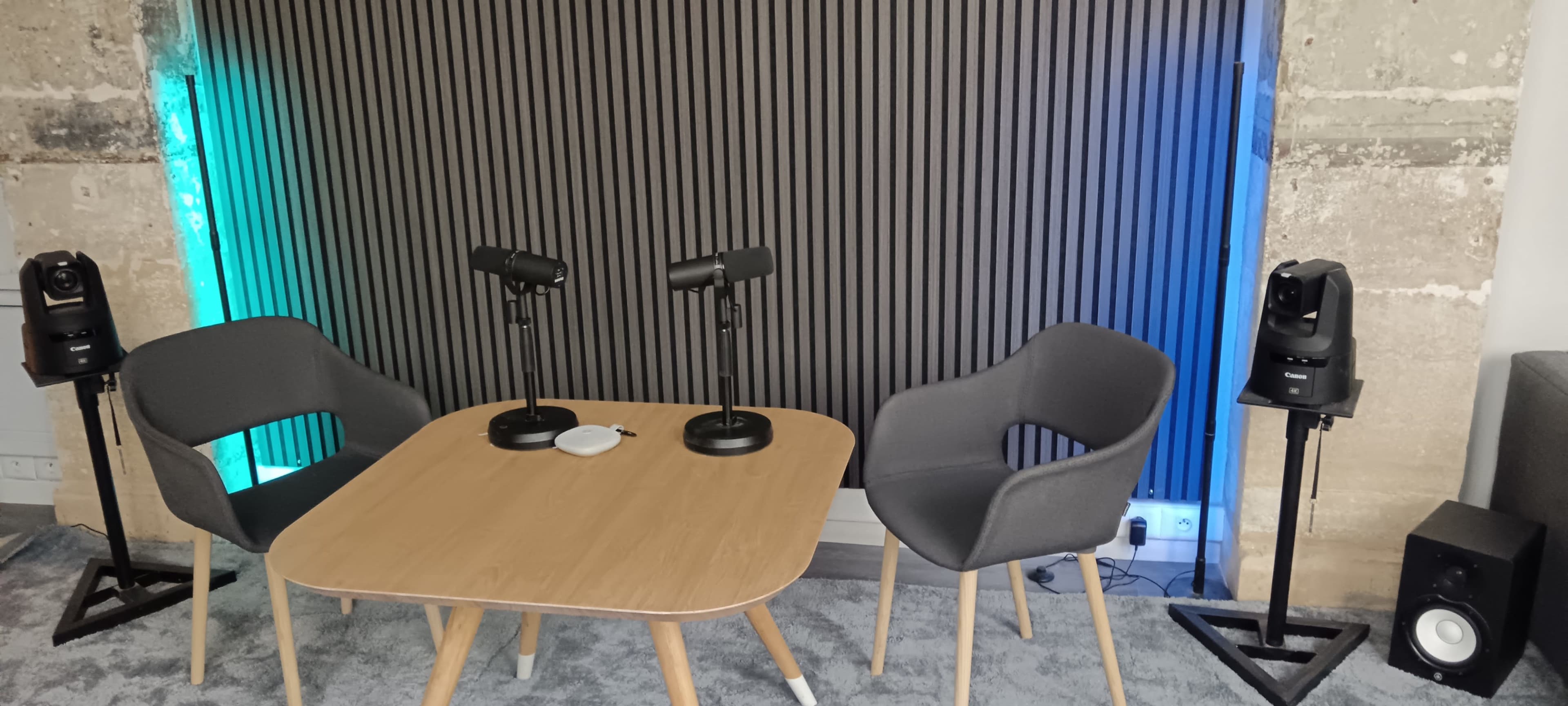 The image shows a modern discussion setup with two microphones on a table, flanked by two chairs and speakers against a textured wall with blue lighting.