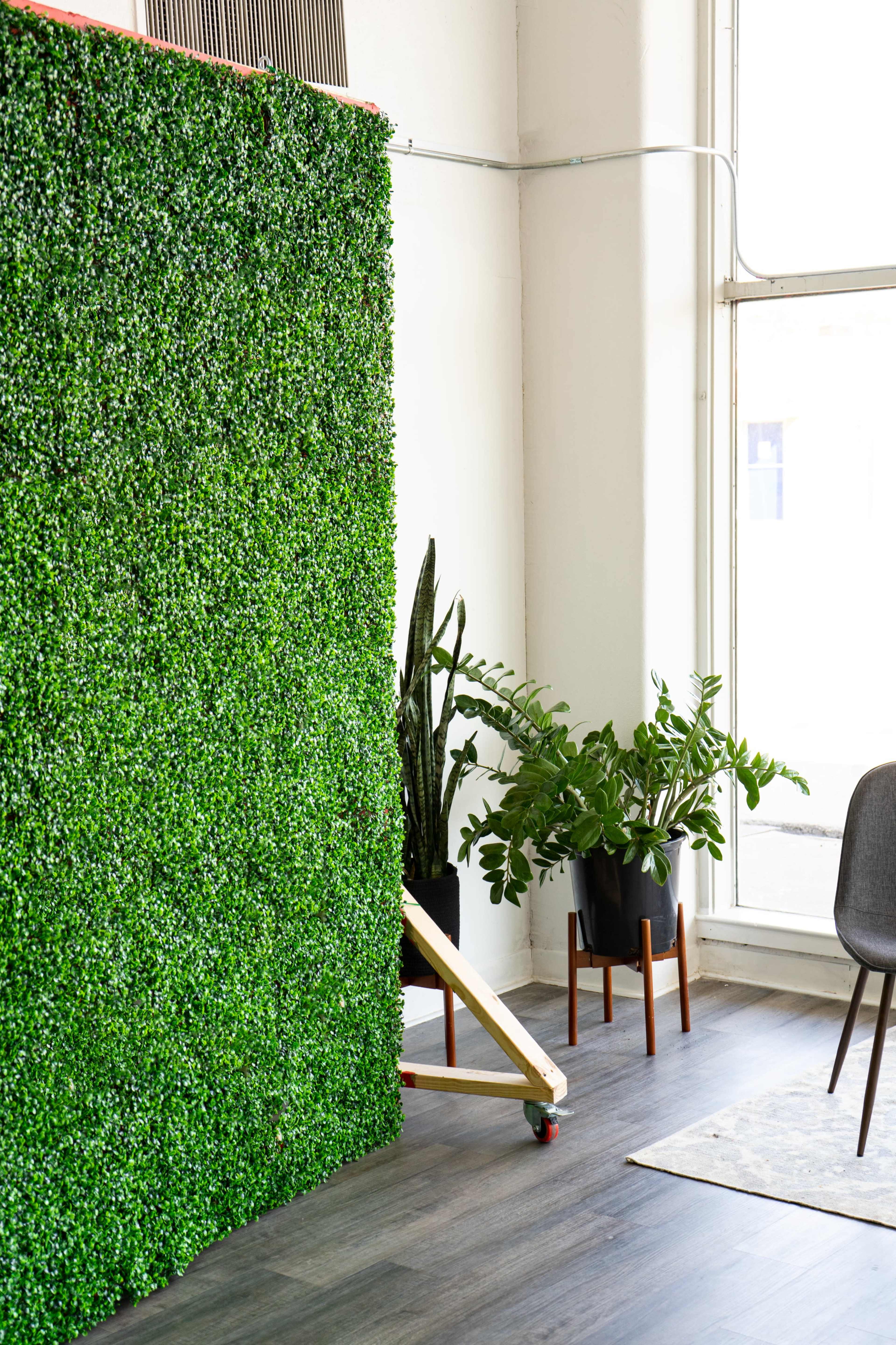 The image shows a room featuring a large artificial green wall, a potted plant, a chair, and a wooden cart.