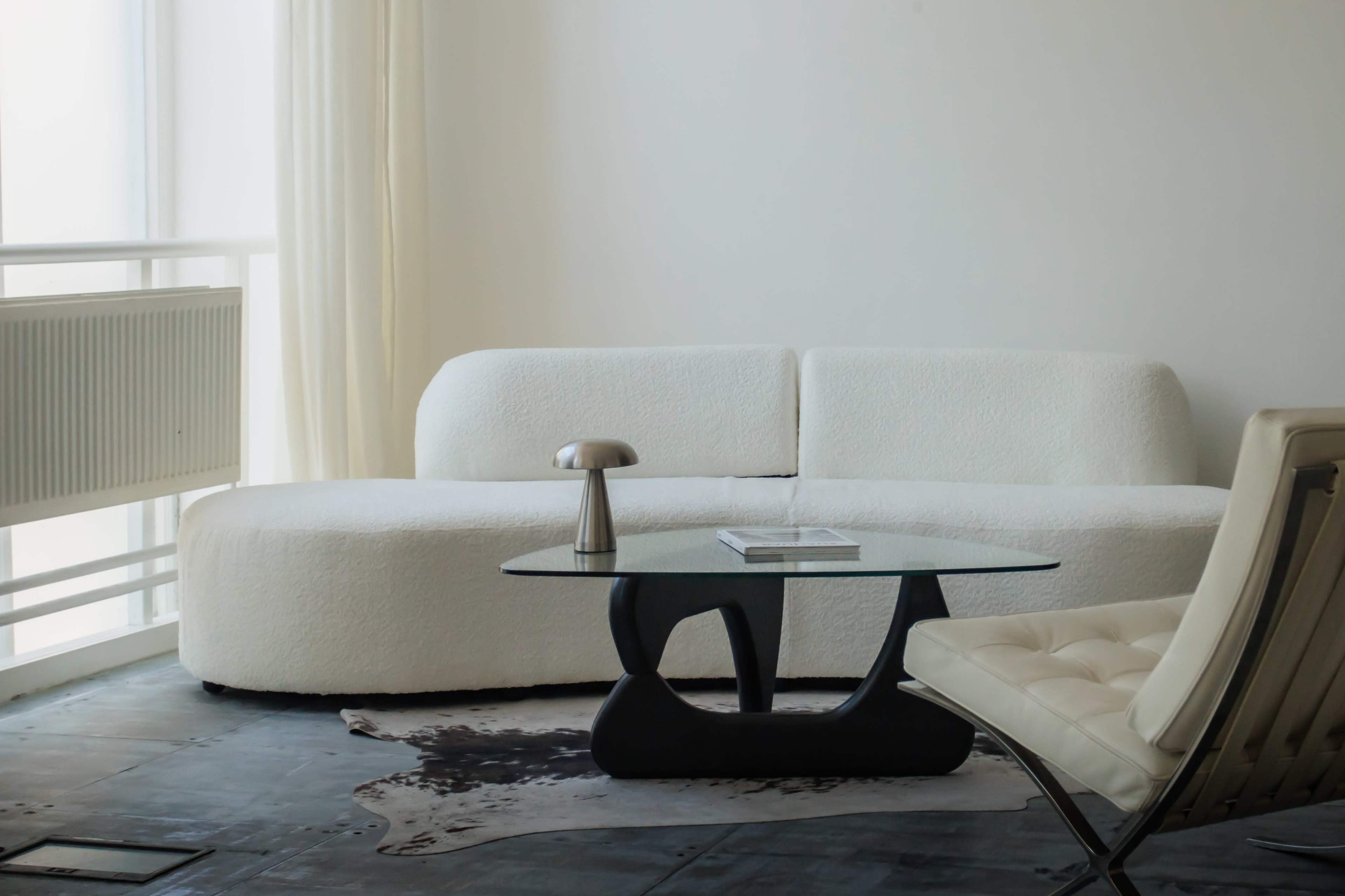 A minimalist living room features a curved white sofa, a sleek black coffee table, and a light-colored chair beside a large window.