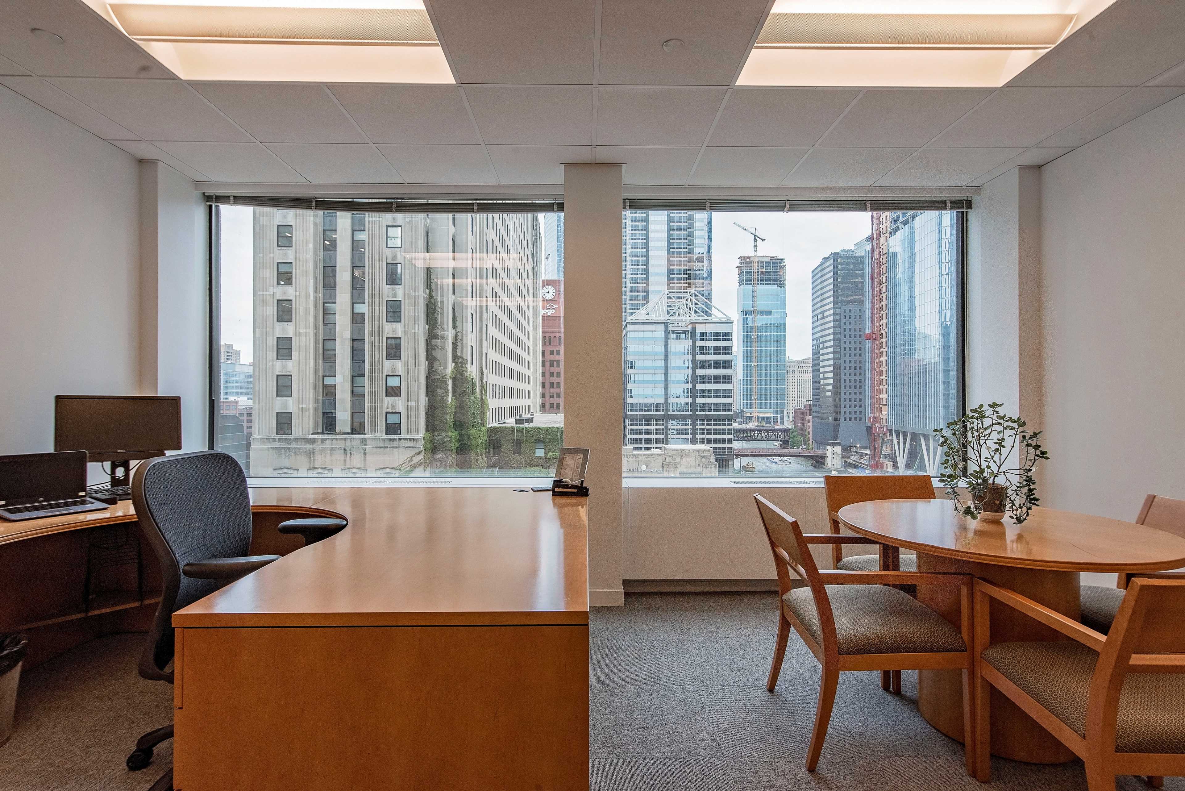 The image shows a modern office space featuring a wooden desk, a black office chair, and a round table with chairs, all against a backdrop of large windows revealing a city skyline.