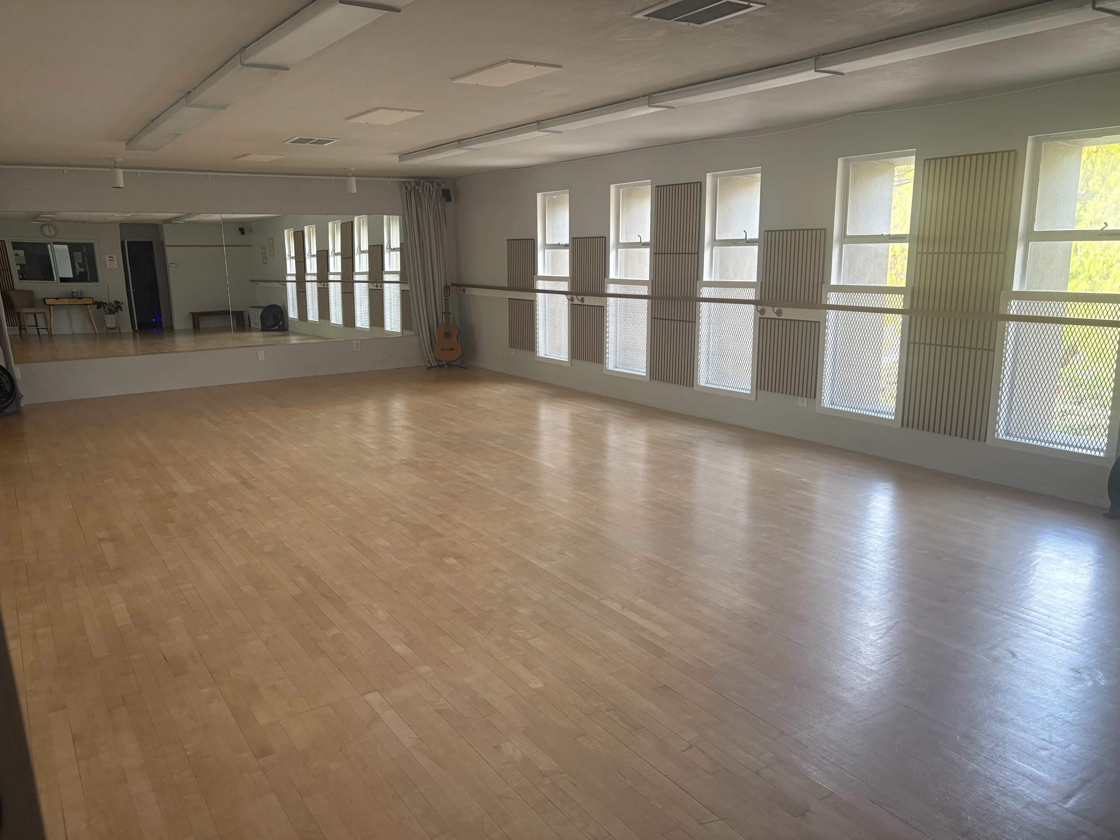 The image shows a spacious dance studio with a wooden floor, large windows, and mirrors along one wall.
