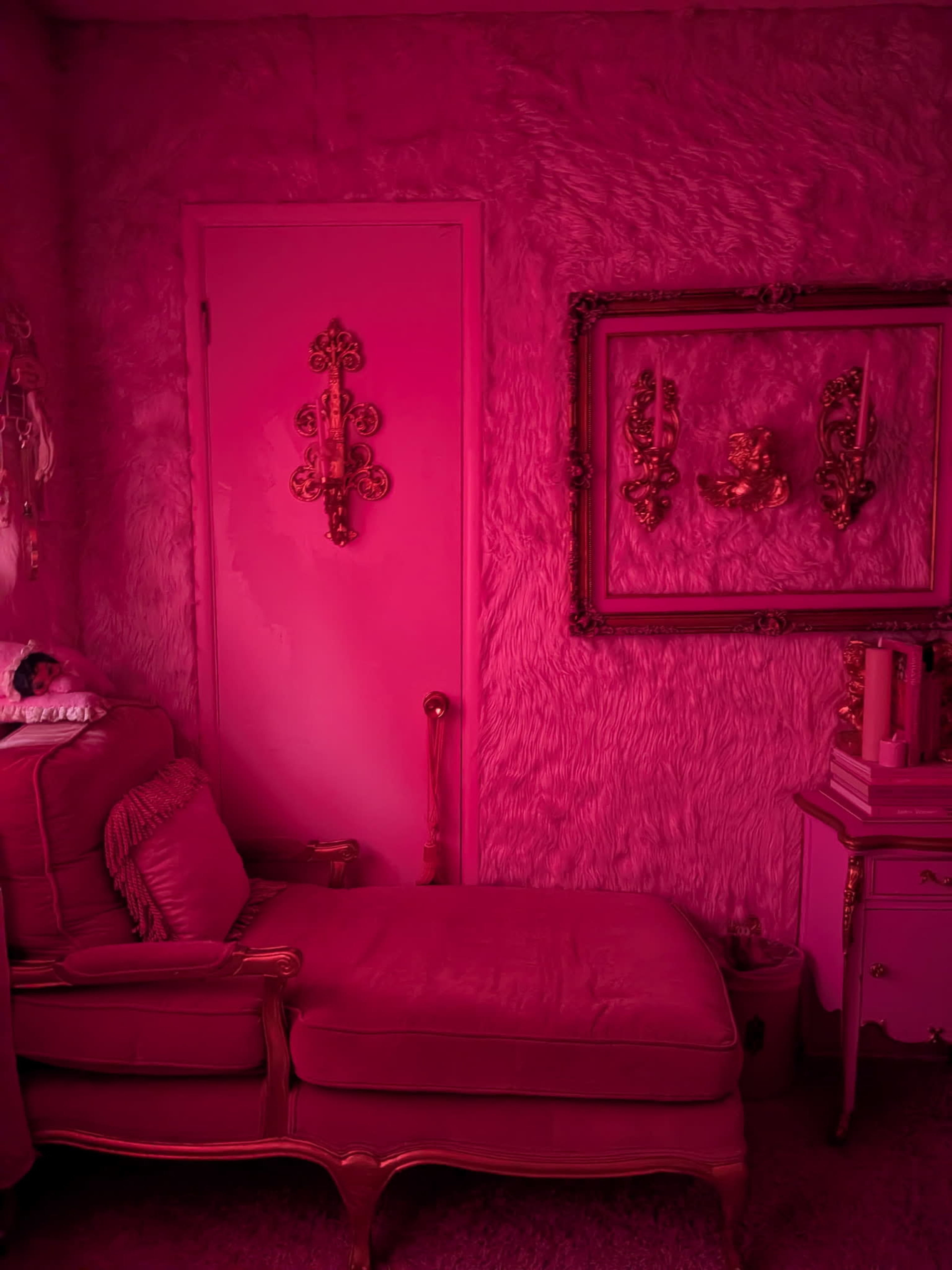 The image shows a pink-themed room featuring a plush chaise lounge, a door, and wall decorations including framed mirrors.