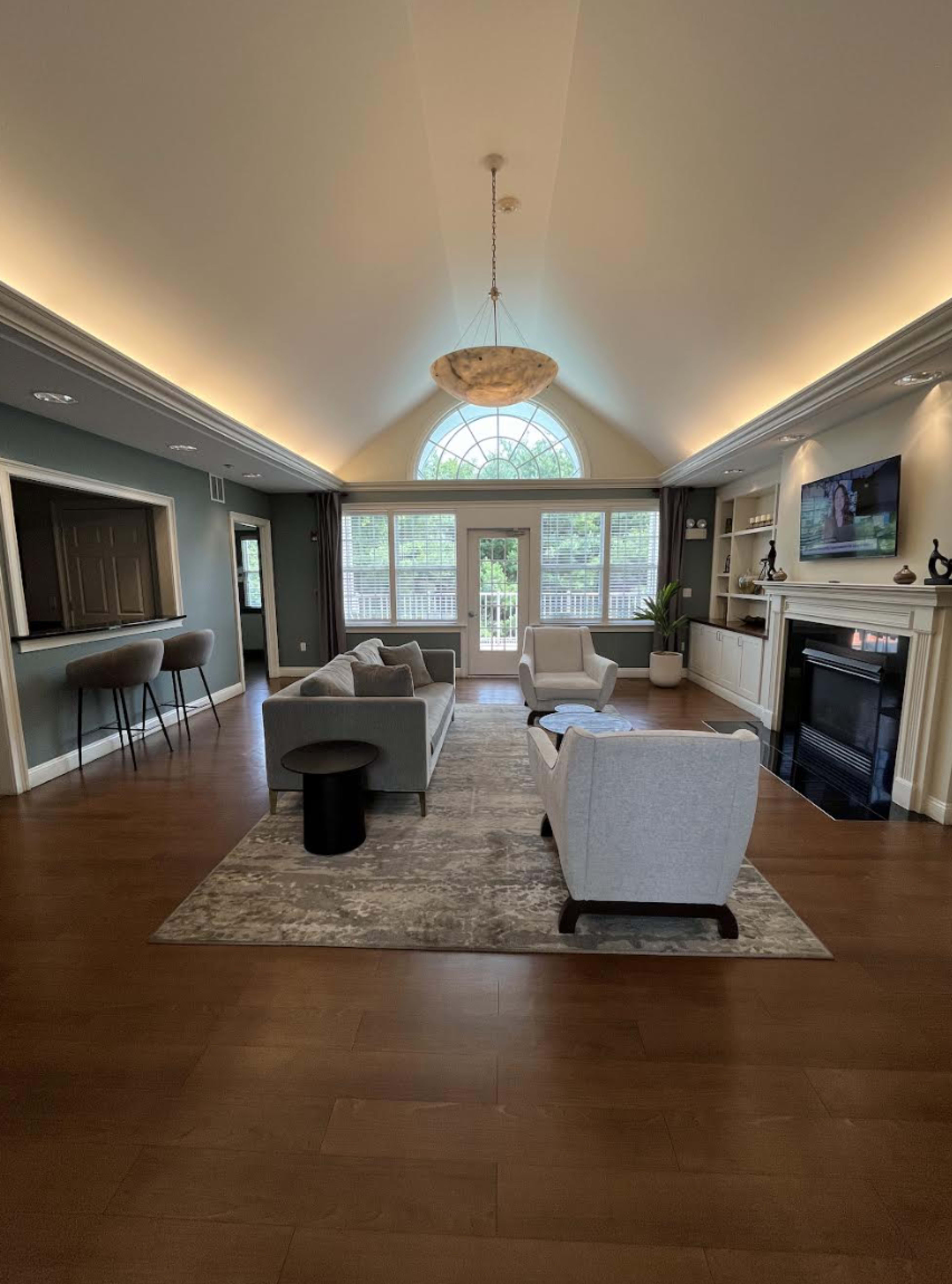 The image shows a spacious living room with two light gray armchairs, a gray sofa, a black side table, a fireplace, and large windows allowing natural light to illuminate the space.