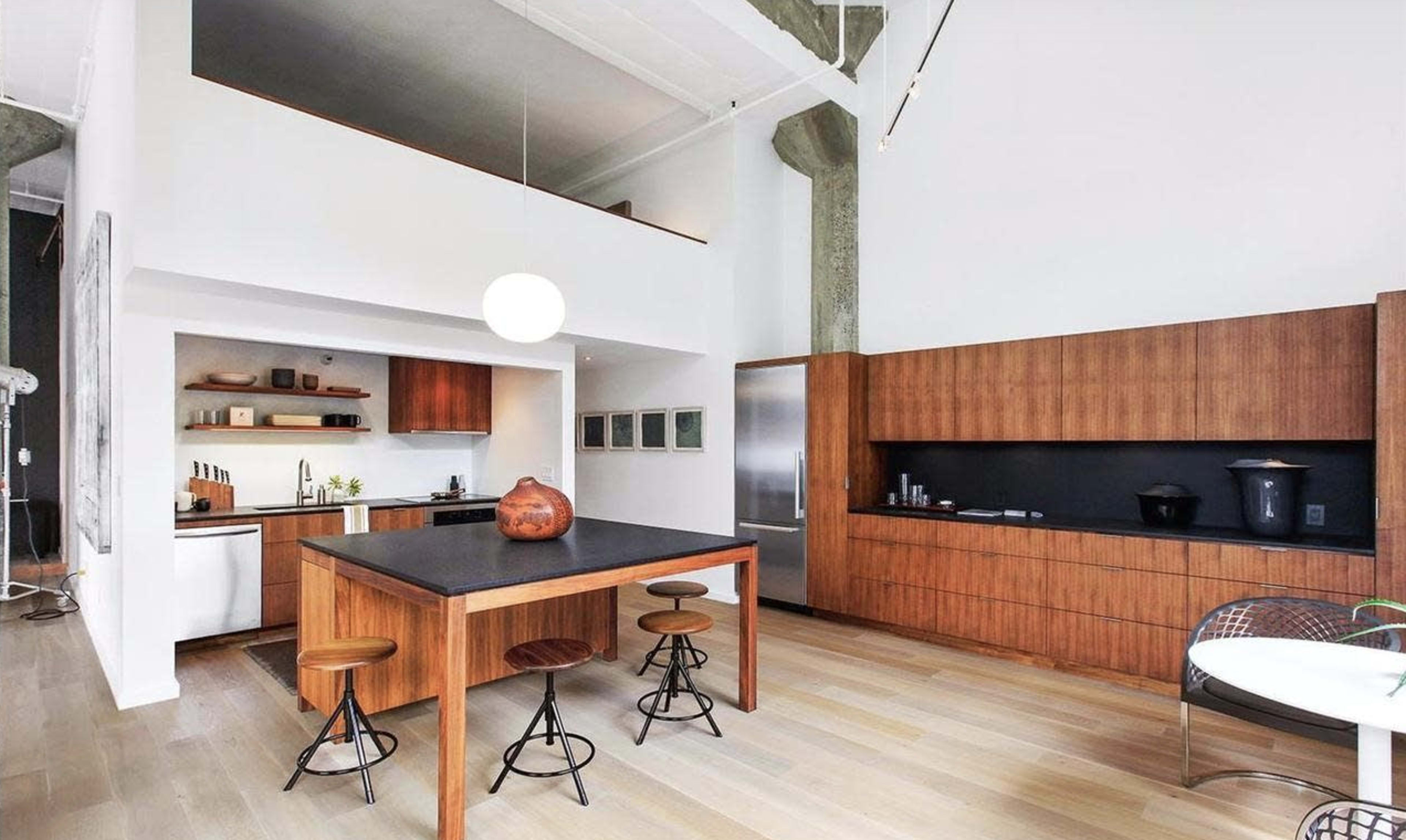 A modern kitchen featuring a central wooden dining table, bar stools, and a minimalist design with wooden cabinetry and a white wall.