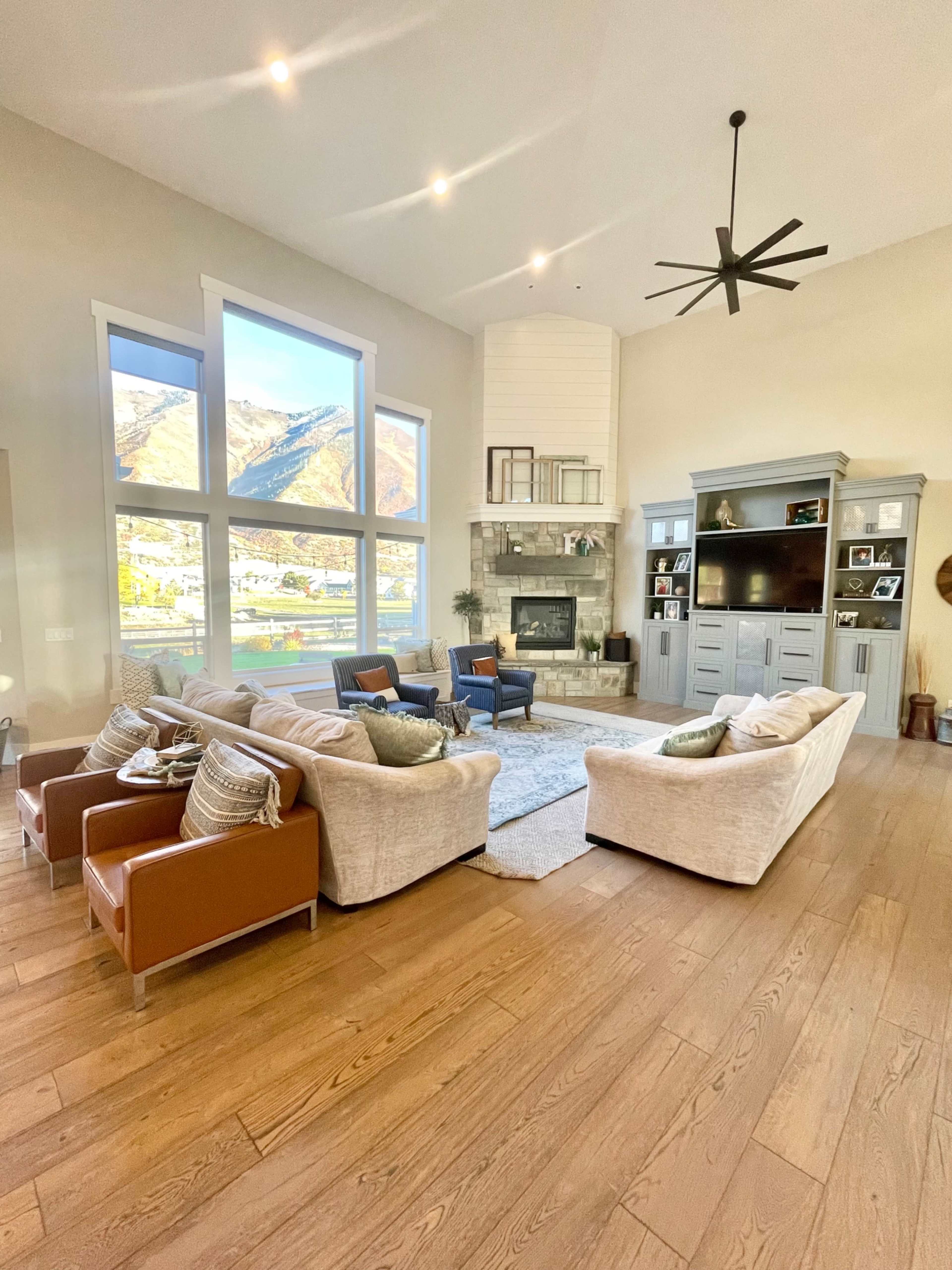 The living room features large windows overlooking a mountainous landscape, a stone fireplace, and seating arrangements comprising two couches and two armchairs.