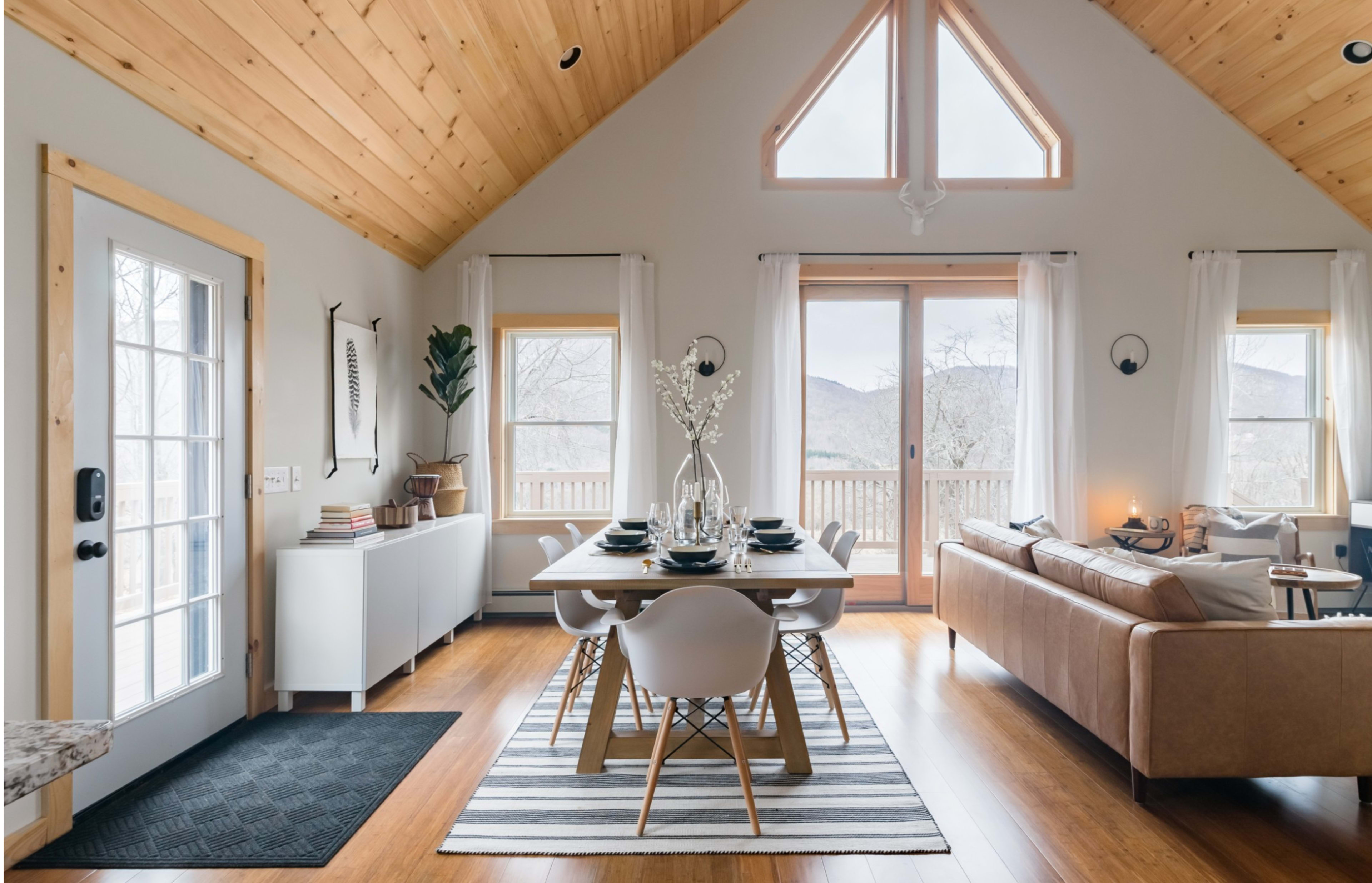 A modern living space features a dining area with a table set for six, a cozy sofa, and large windows overlooking a mountain view.