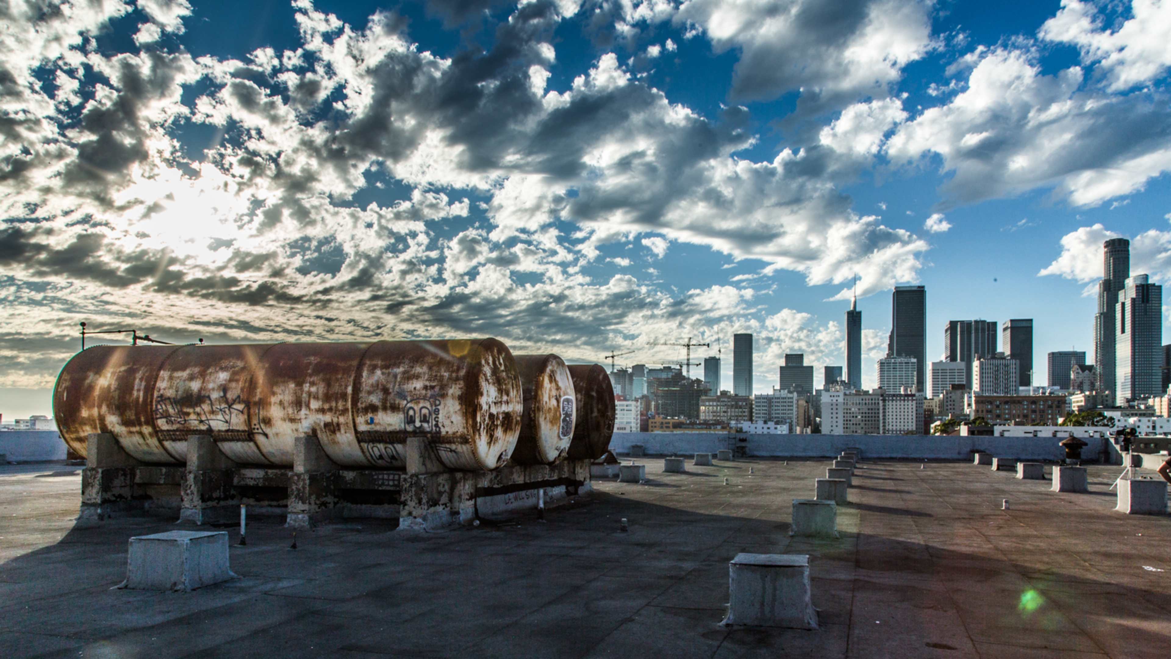 Rusty storage tanks on a rooftop with a skyline of tall buildings and a dramatic sky filled with clouds in the background.