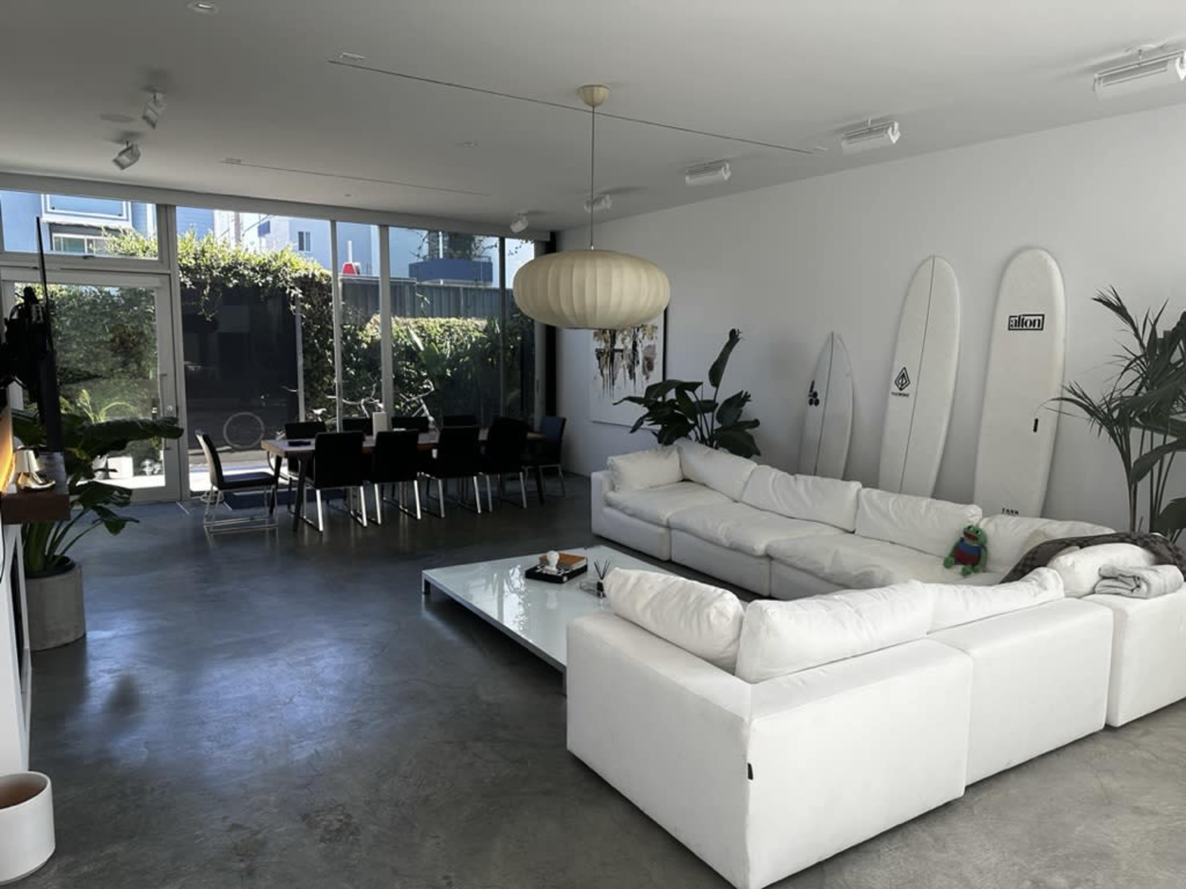 A modern living room with a large white sectional sofa, a glass coffee table, potted plants, and surfboards leaning against the wall, leading to an outdoor dining area visible through large windows.