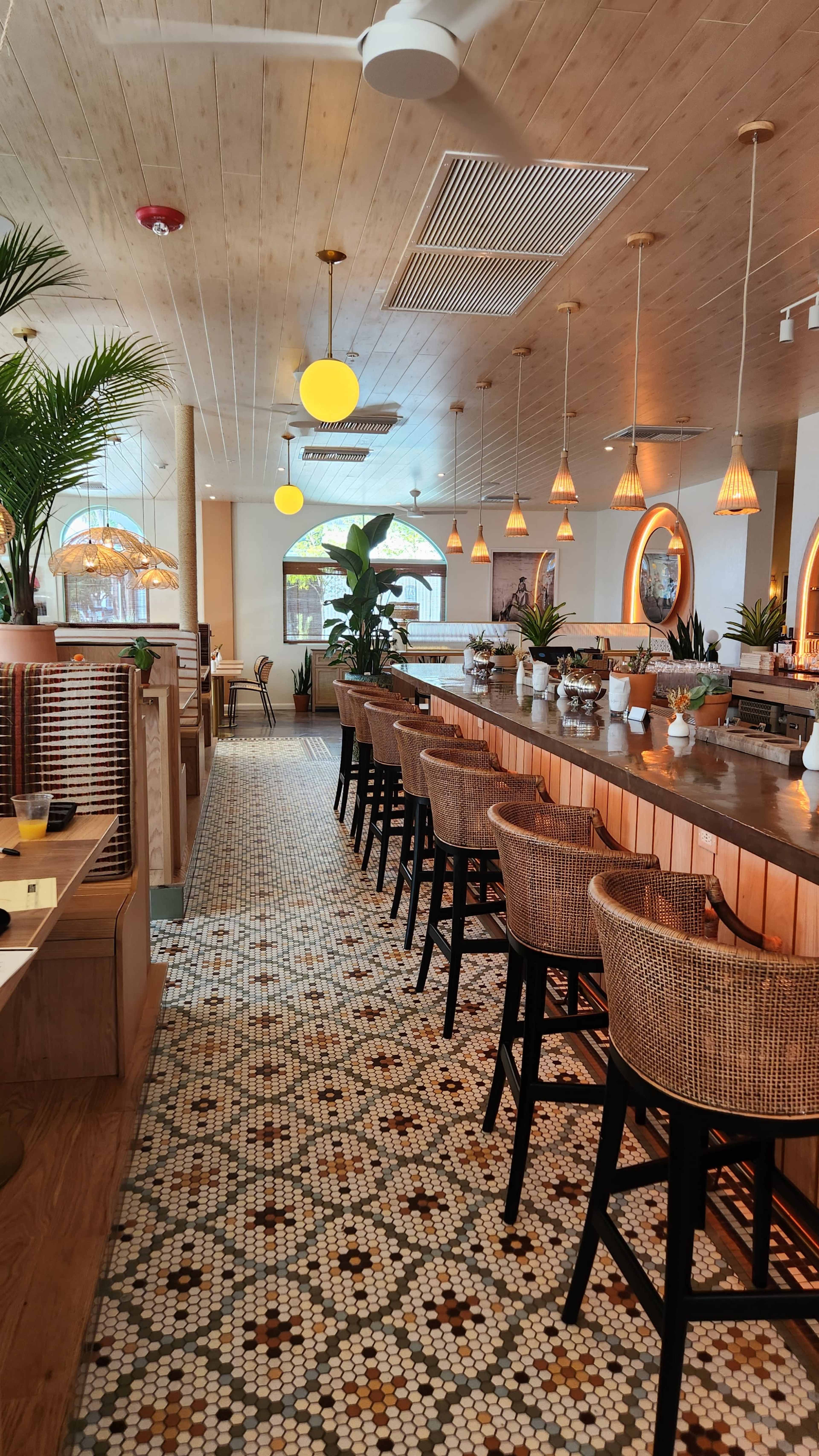 The image shows a spacious, modern restaurant interior featuring a long wooden bar with high stools, decorative lighting, and tiled flooring with a pattern.