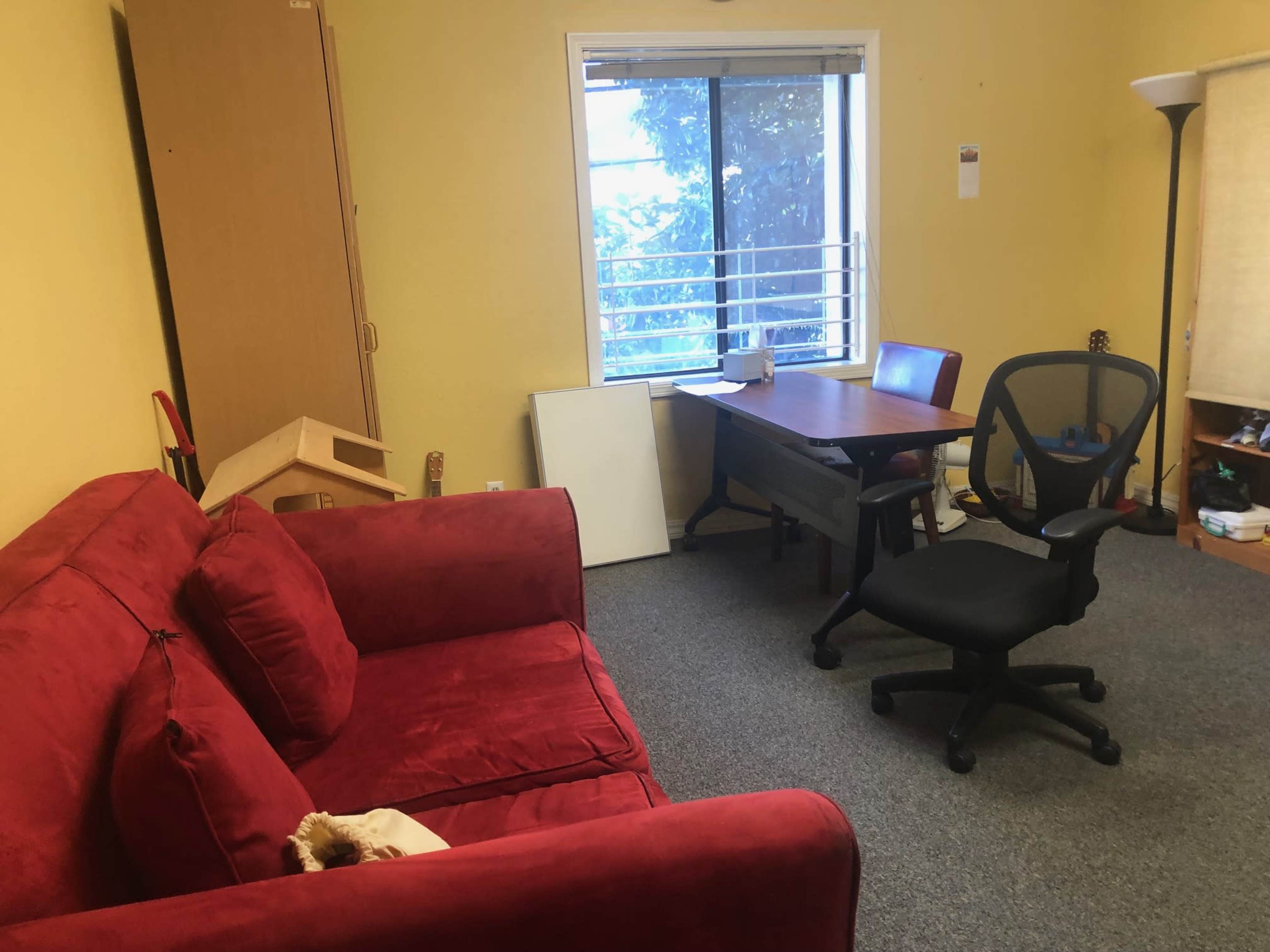 The image shows a room with a red couch, a desk with a chair, and a window letting in natural light.