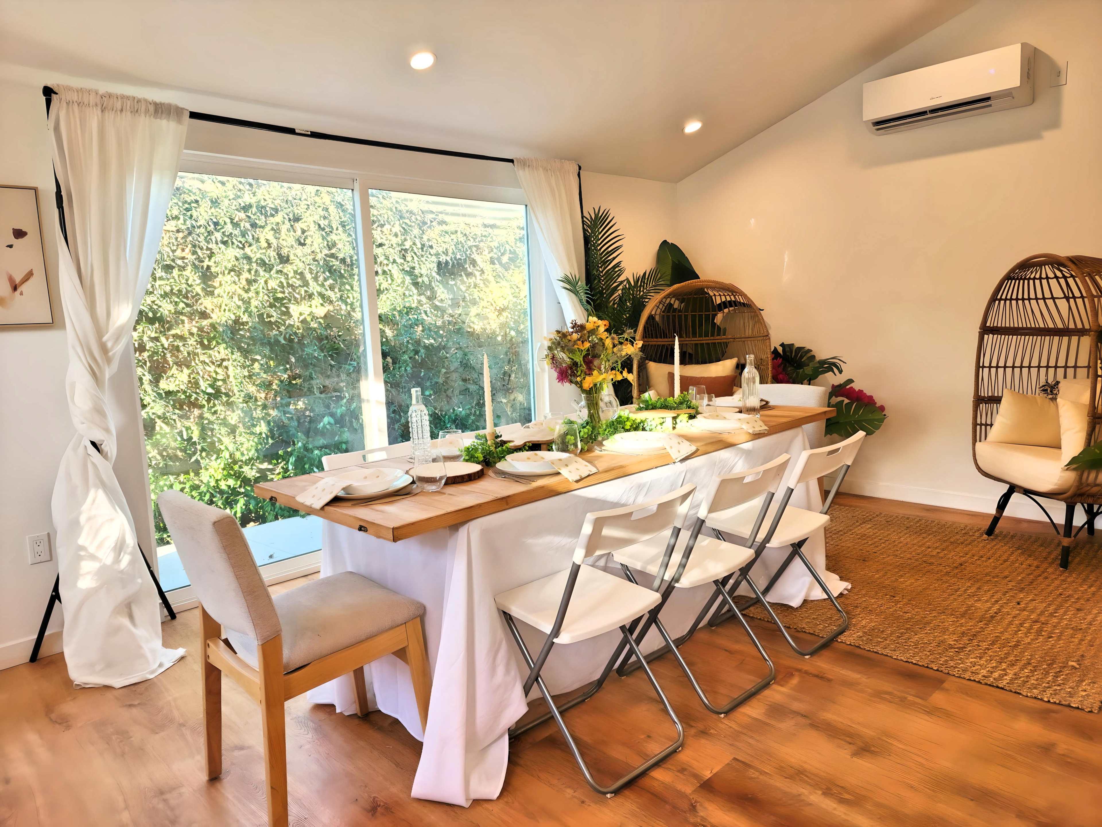 A long dining table is set with plates and glasses, surrounded by folding chairs, with large windows opening to a lush garden.