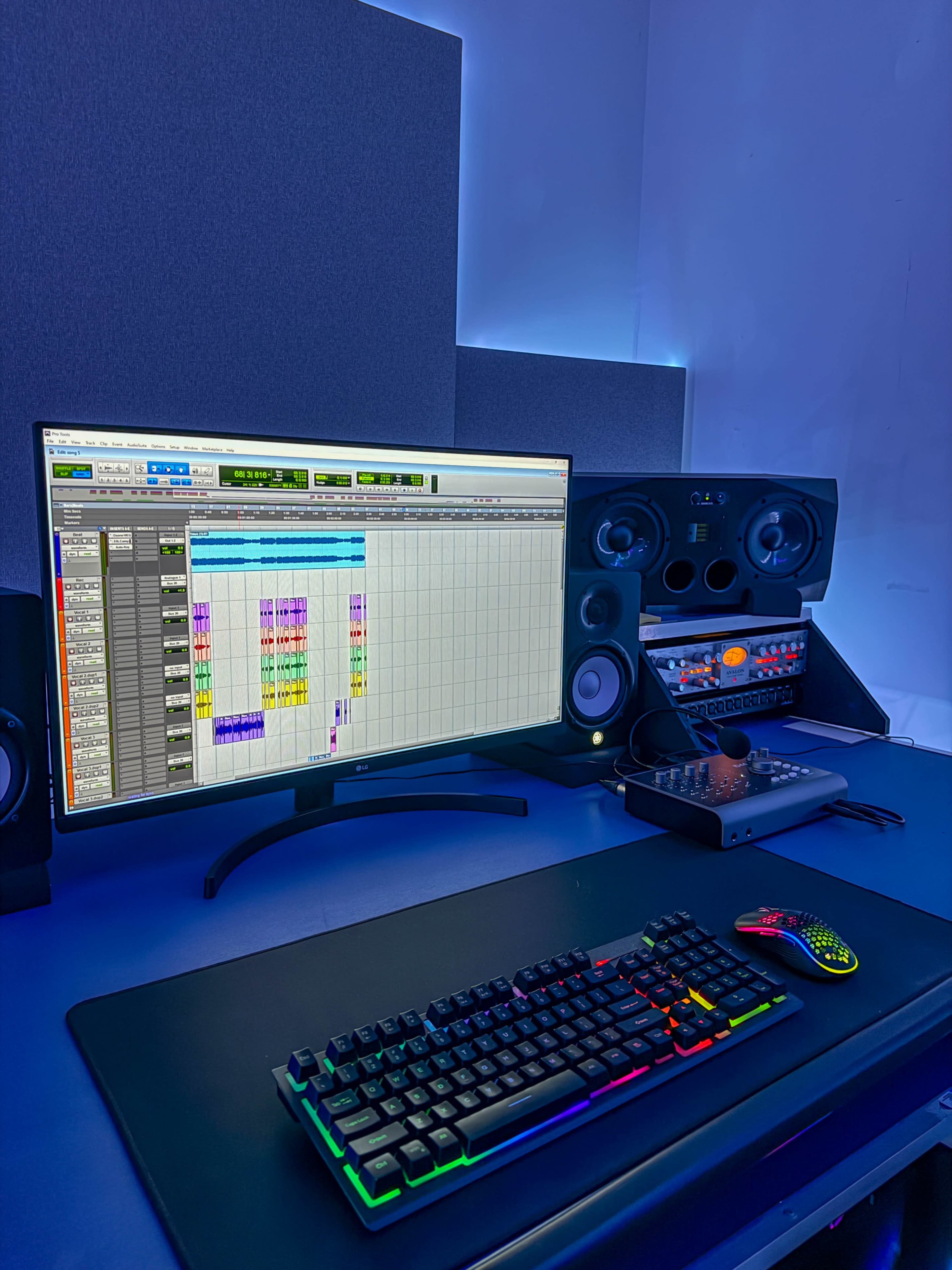 A modern music production setup features a large monitor displaying audio editing software, a keyboard with colorful lighting, studio monitors, and a digital audio interface on a sleek desk.