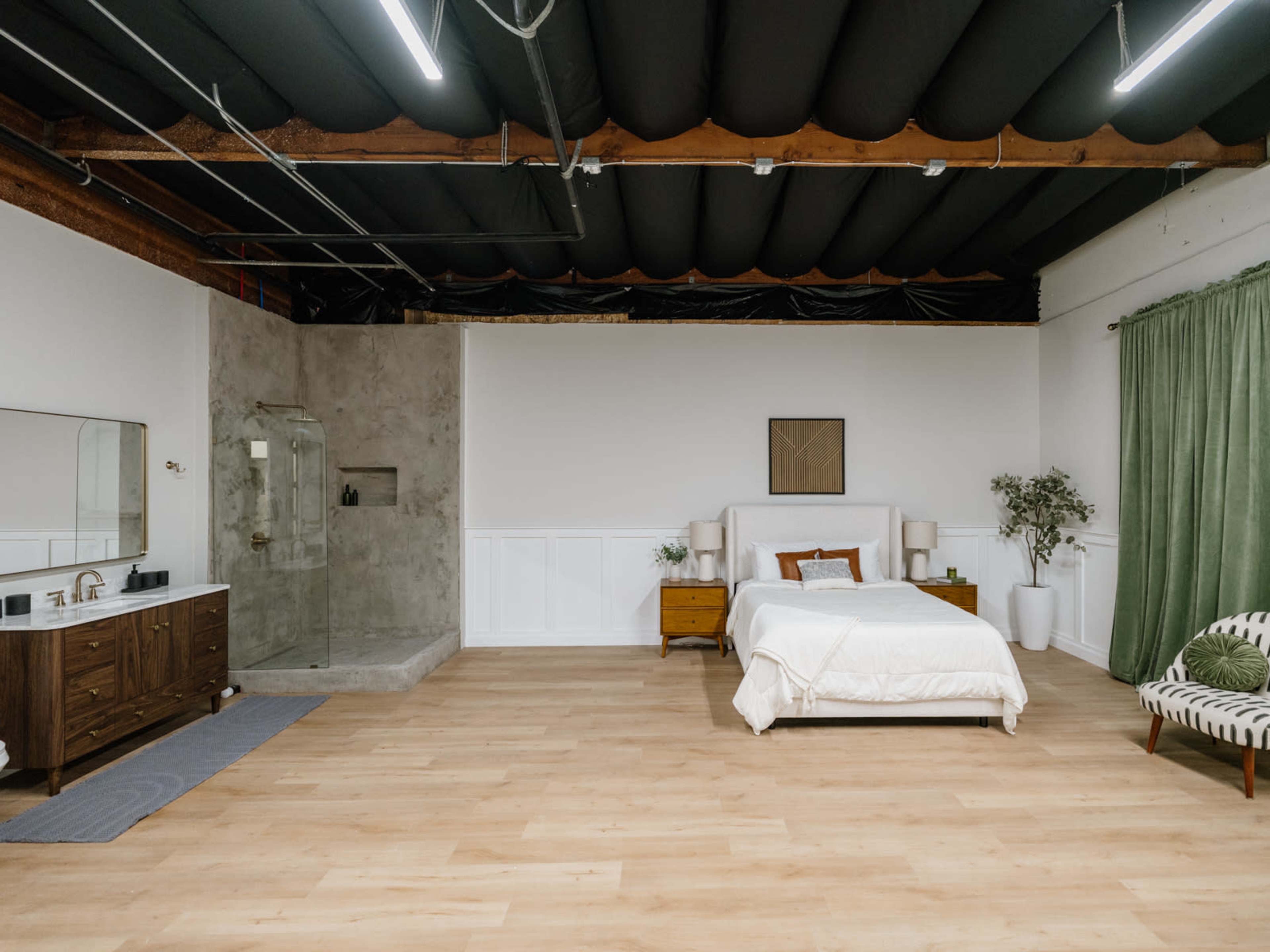The image shows a spacious bedroom with a bed, a wooden dresser, a bathroom area featuring a glass shower, and large windows covered with green curtains.