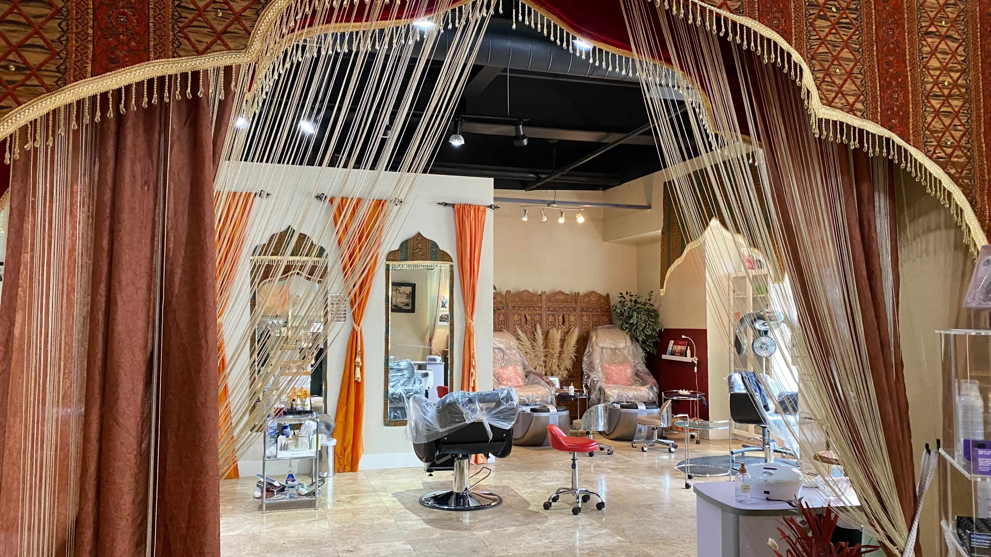 A salon interior features hair styling stations separated by decorative fabric drapes, with a warm color palette and ornate walls.