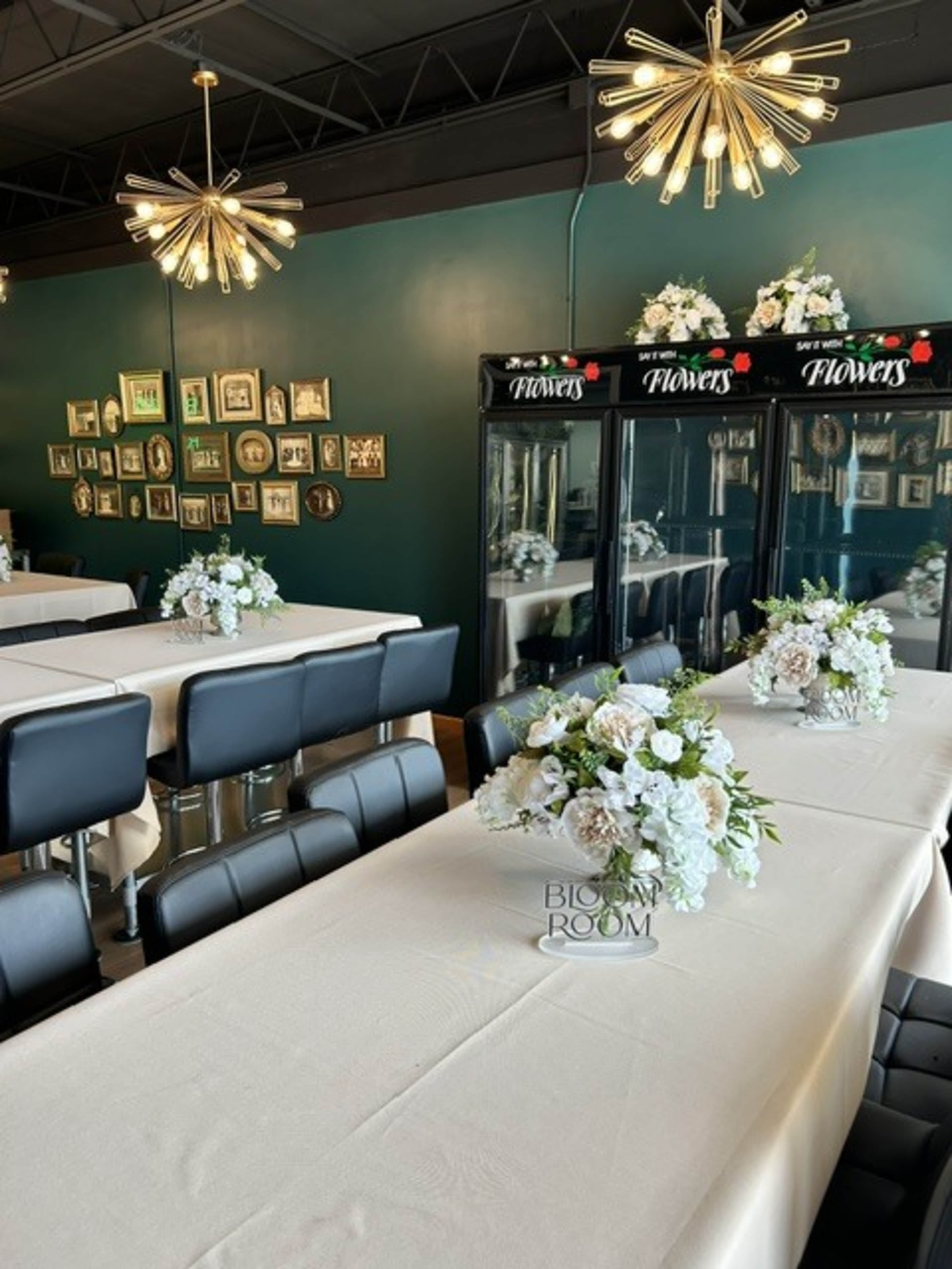 The image shows a dining area with long tables covered in beige tablecloths, adorned with floral centerpieces, and featuring a wall decorated with framed pictures and floral displays in glass cases.