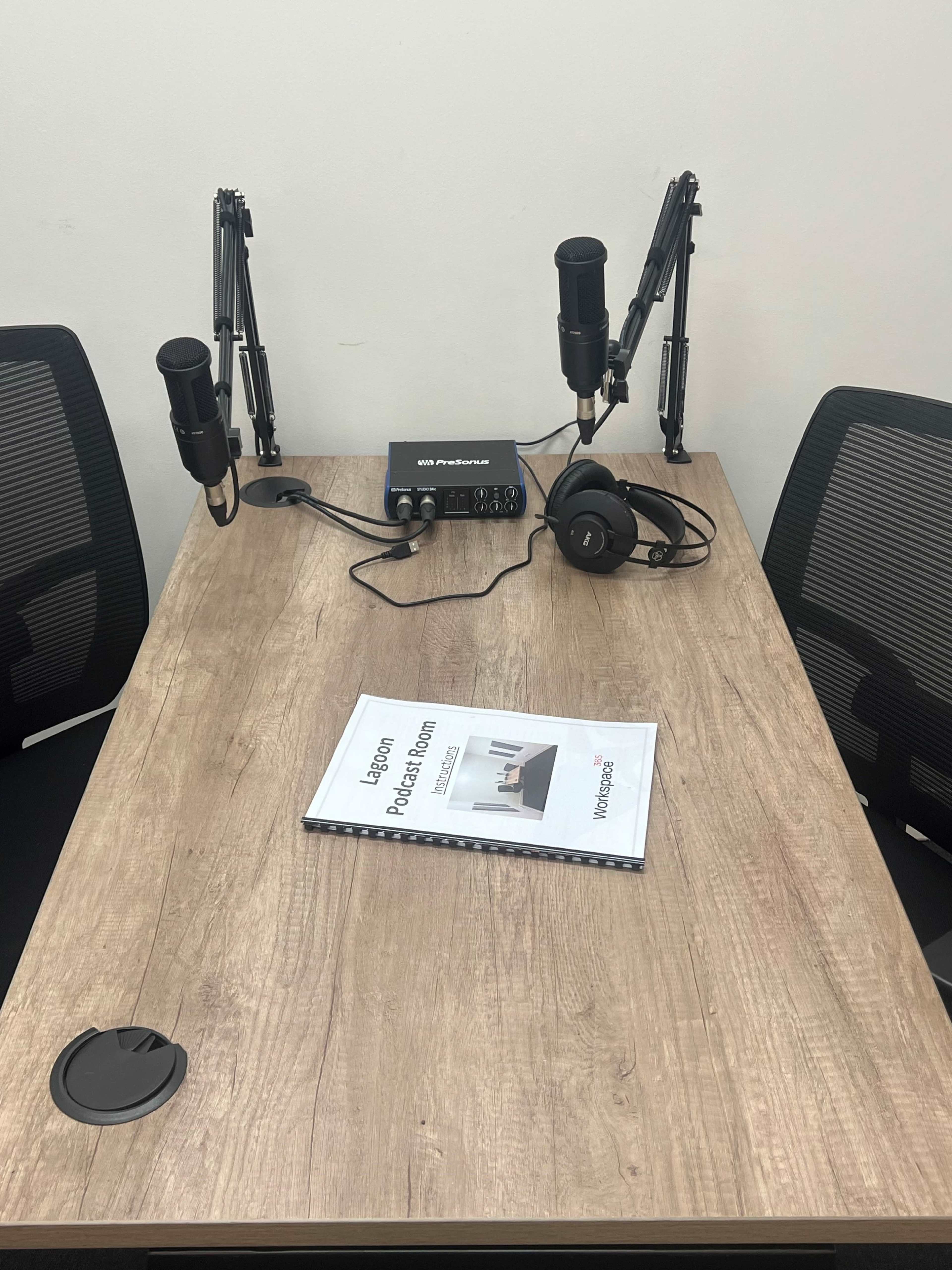 The image shows a podcast recording setup featuring two microphones, an audio interface, a pair of headphones, and a notebook on a wooden table, with two chairs positioned on either side.