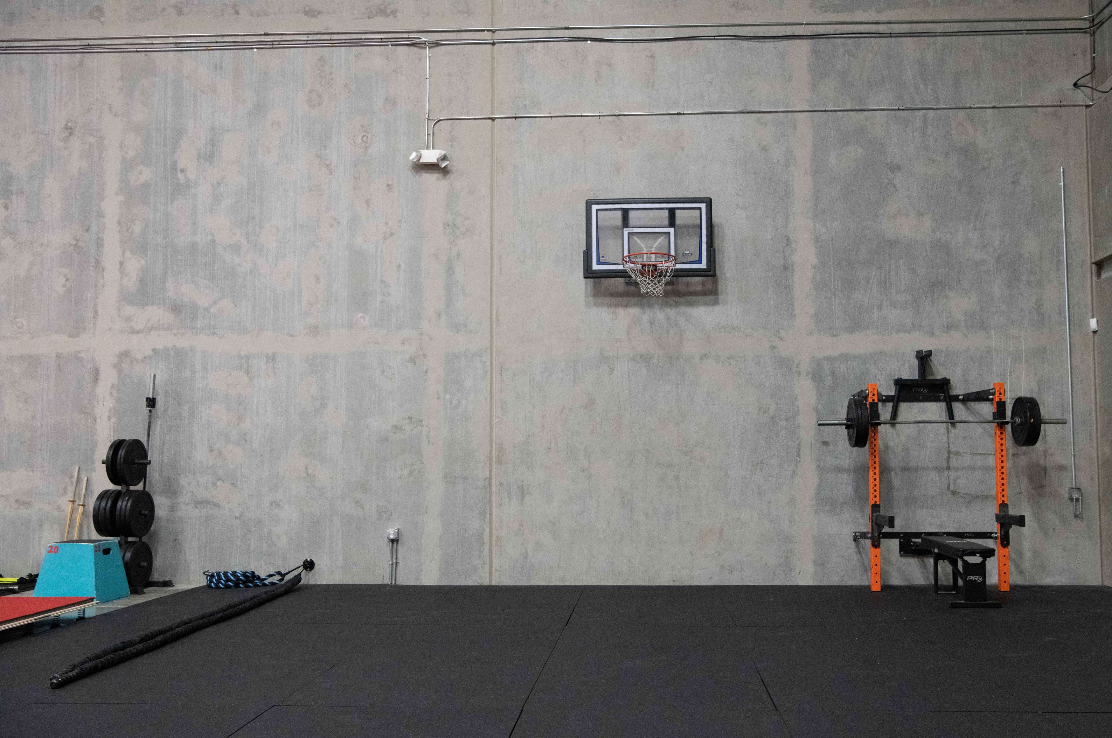 A gym interior featuring a basketball hoop, weightlifting equipment, and exercise mats against a grey concrete wall.