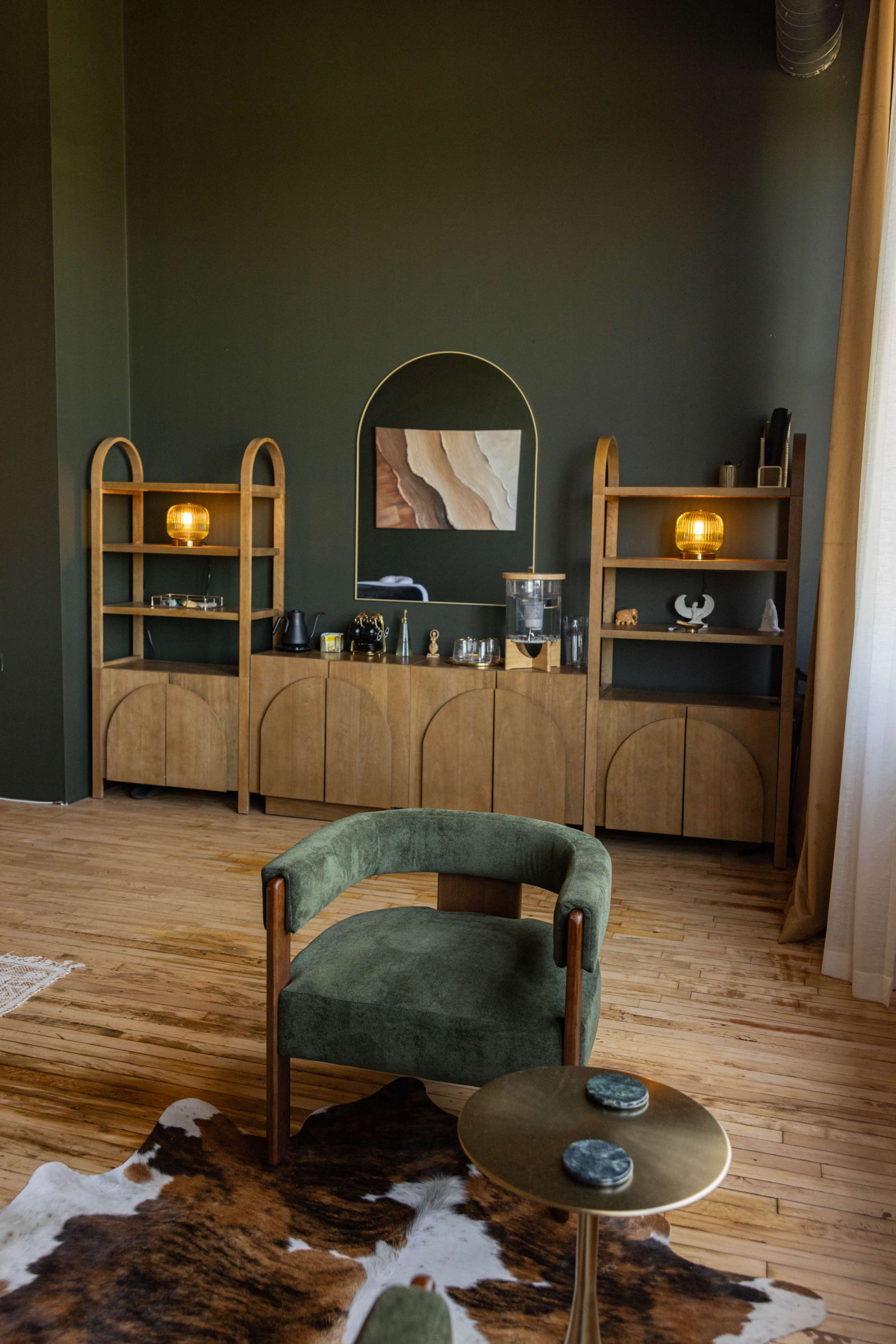 The image features a cozy interior with a green upholstered chair, a wooden shelving unit displaying decorative items, and soft lighting against a dark green wall.