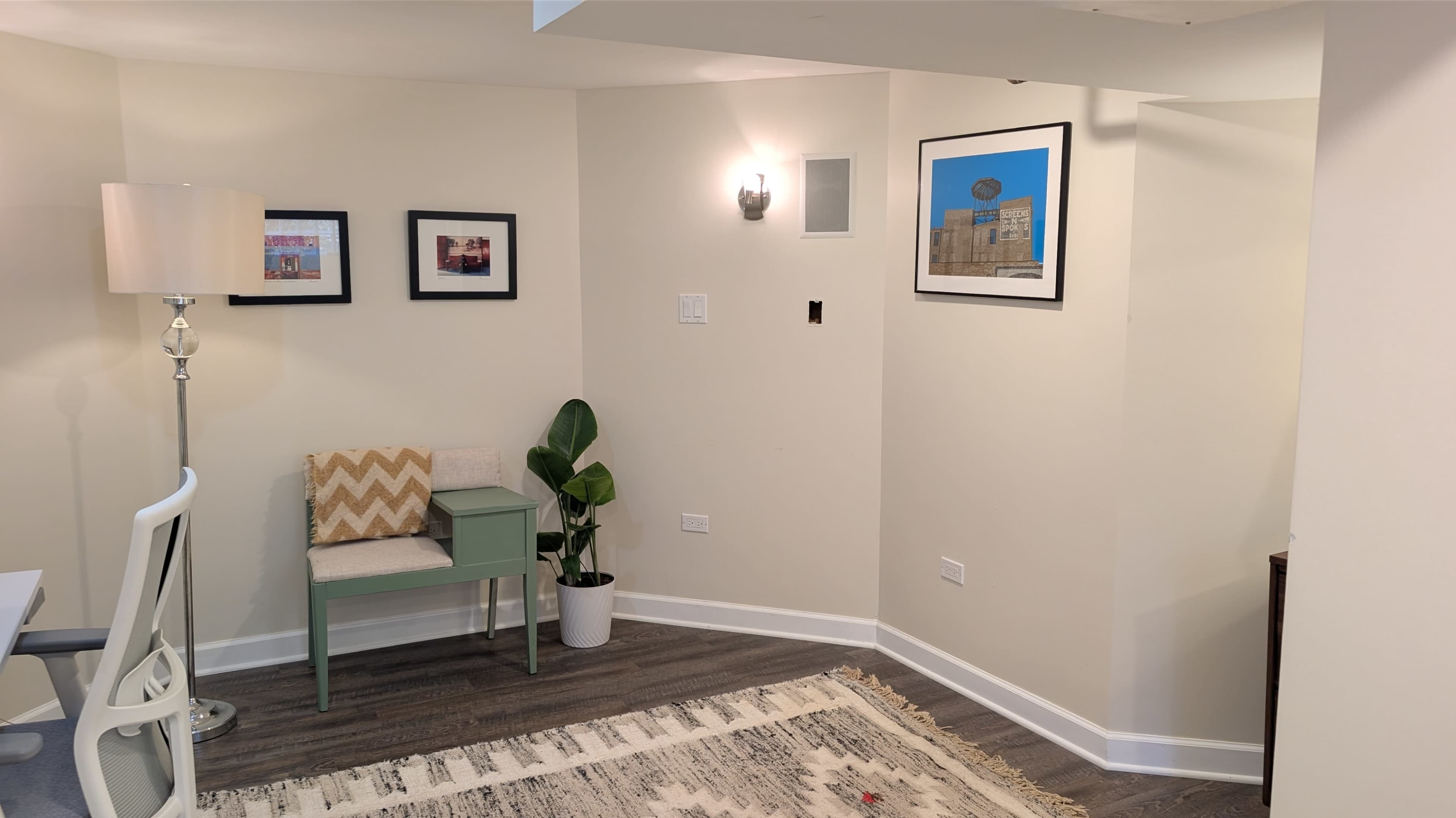 The image shows a small room with a desk and chair, a green accent table, decorative framed pictures on the walls, a lamp, a plant, and a patterned rug on the floor.