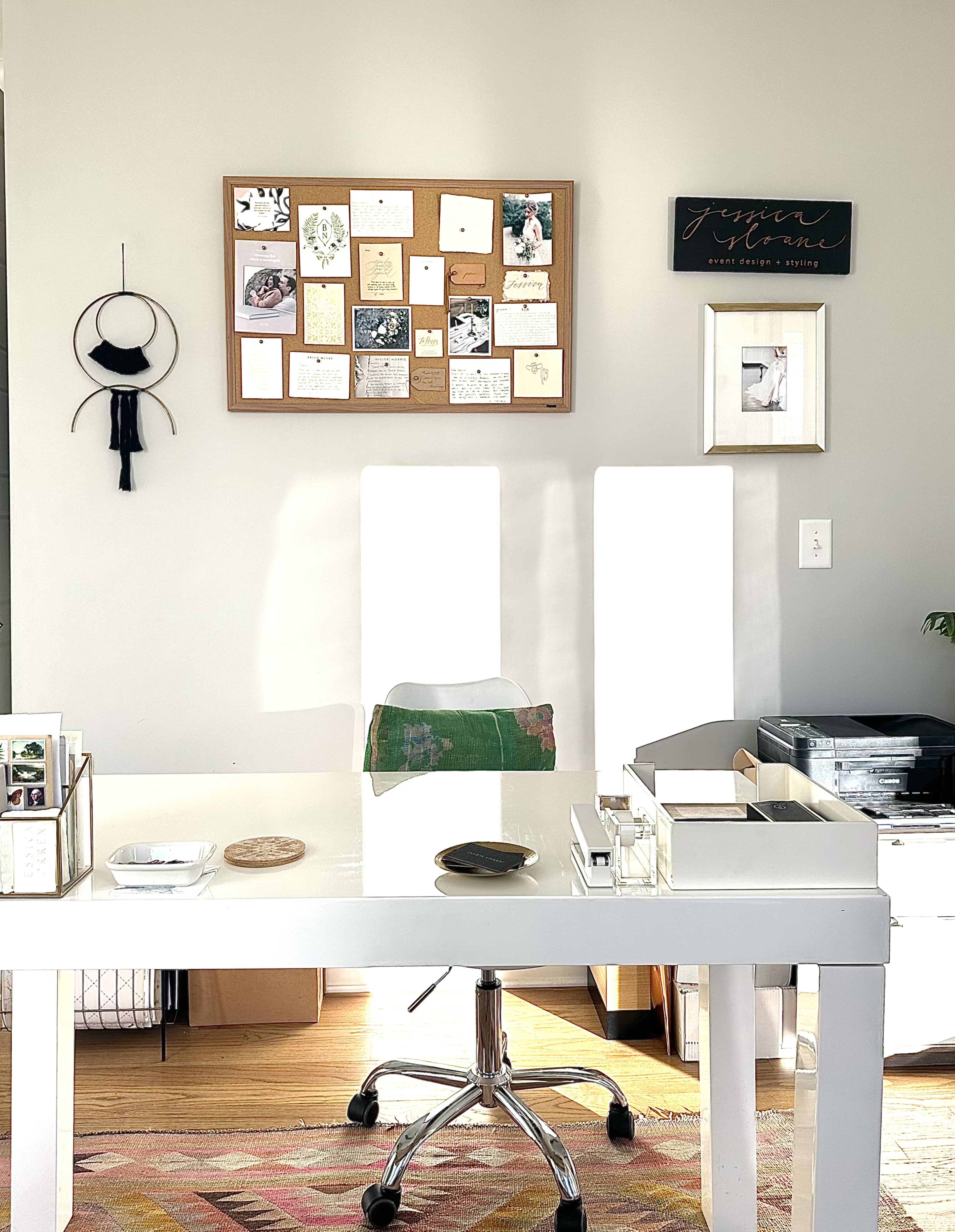 A well-lit home office features a white desk and chair, a bulletin board with various pinned notes and images, and storage boxes in the background.