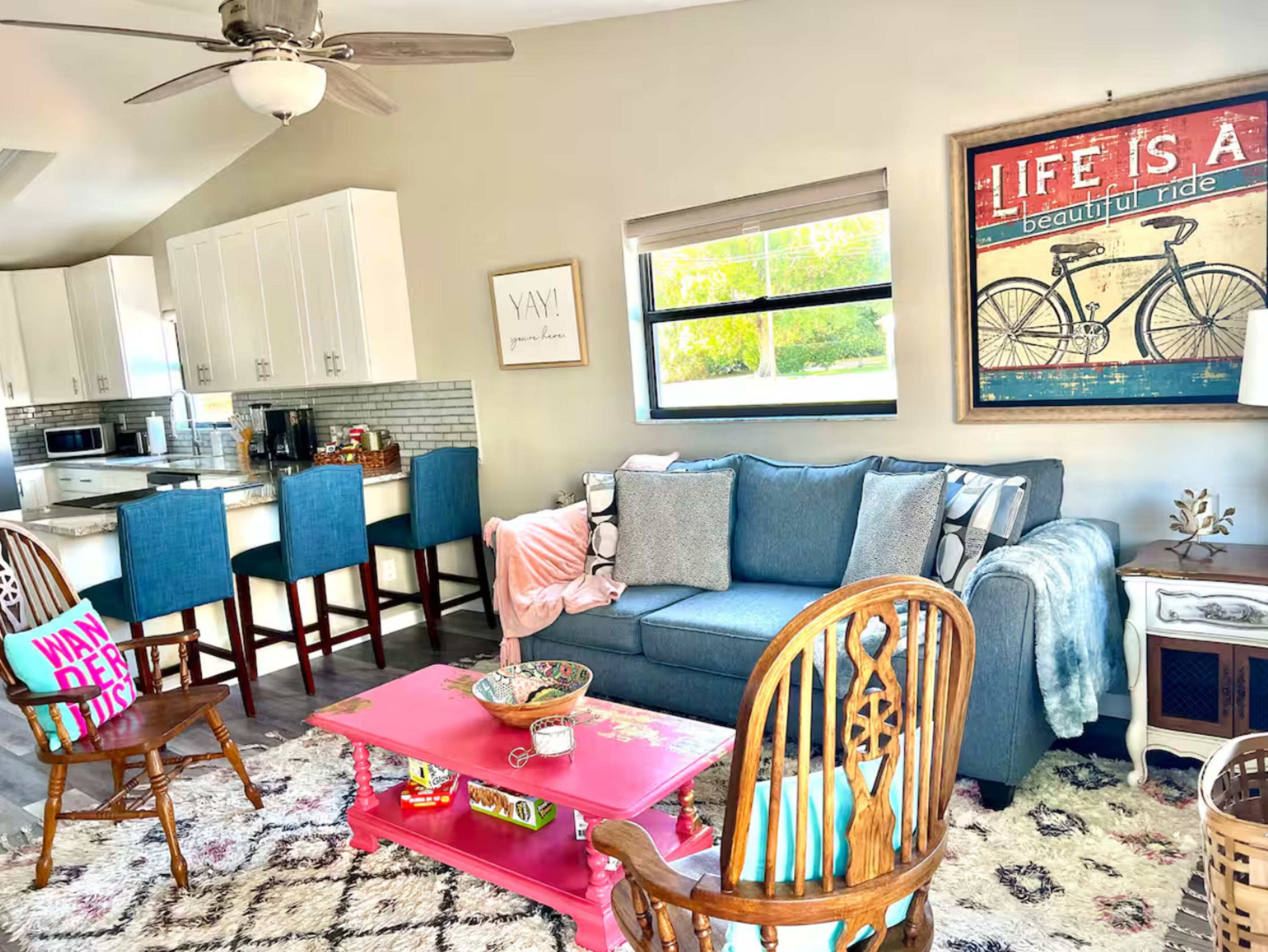 The image shows a living room featuring a blue couch, a pink coffee table, and a kitchen area in the background with white cabinets and bar stools.