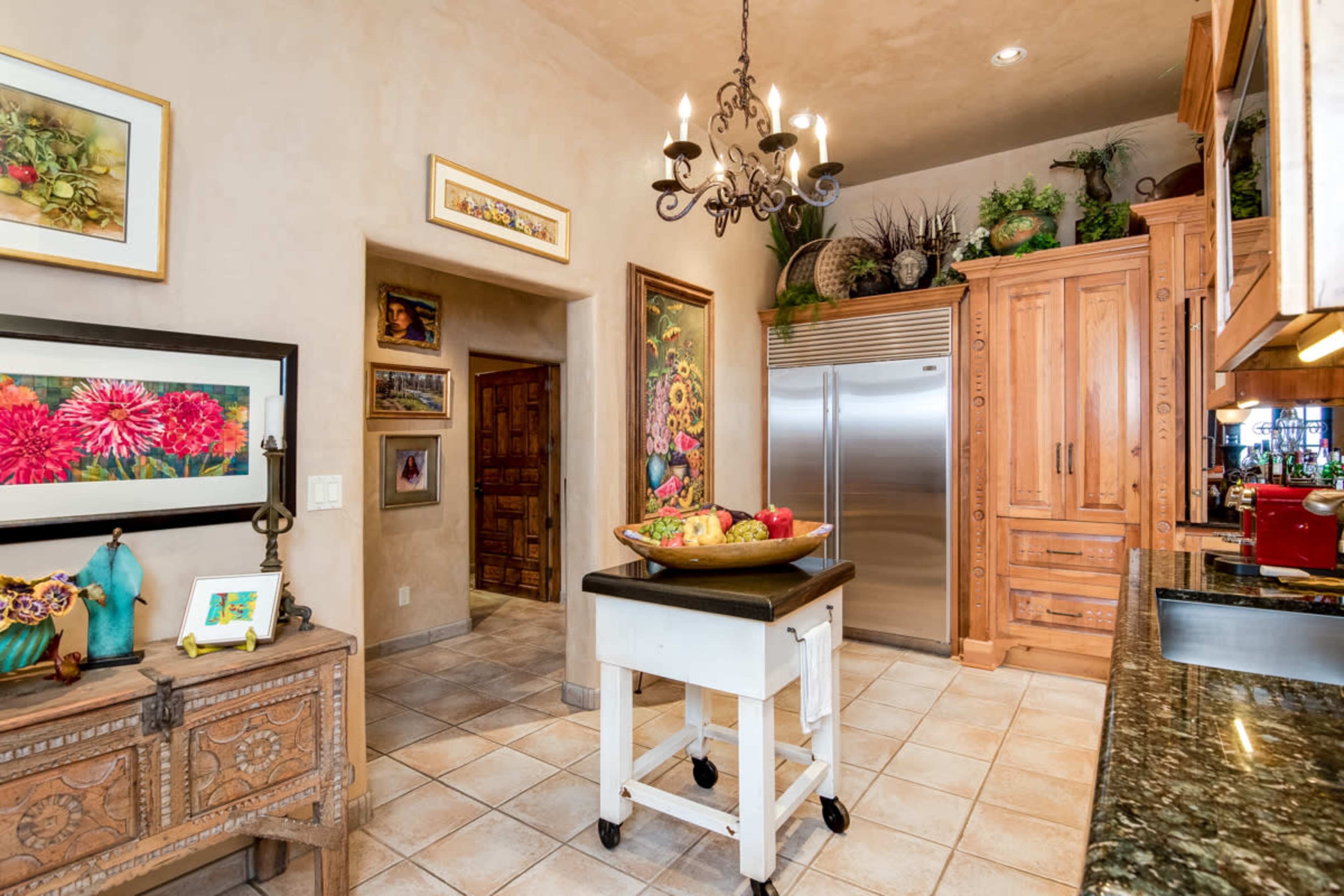 The kitchen features wooden cabinetry, a stainless steel refrigerator, a chandelier, and a decorative fruit display on a rolling cart.