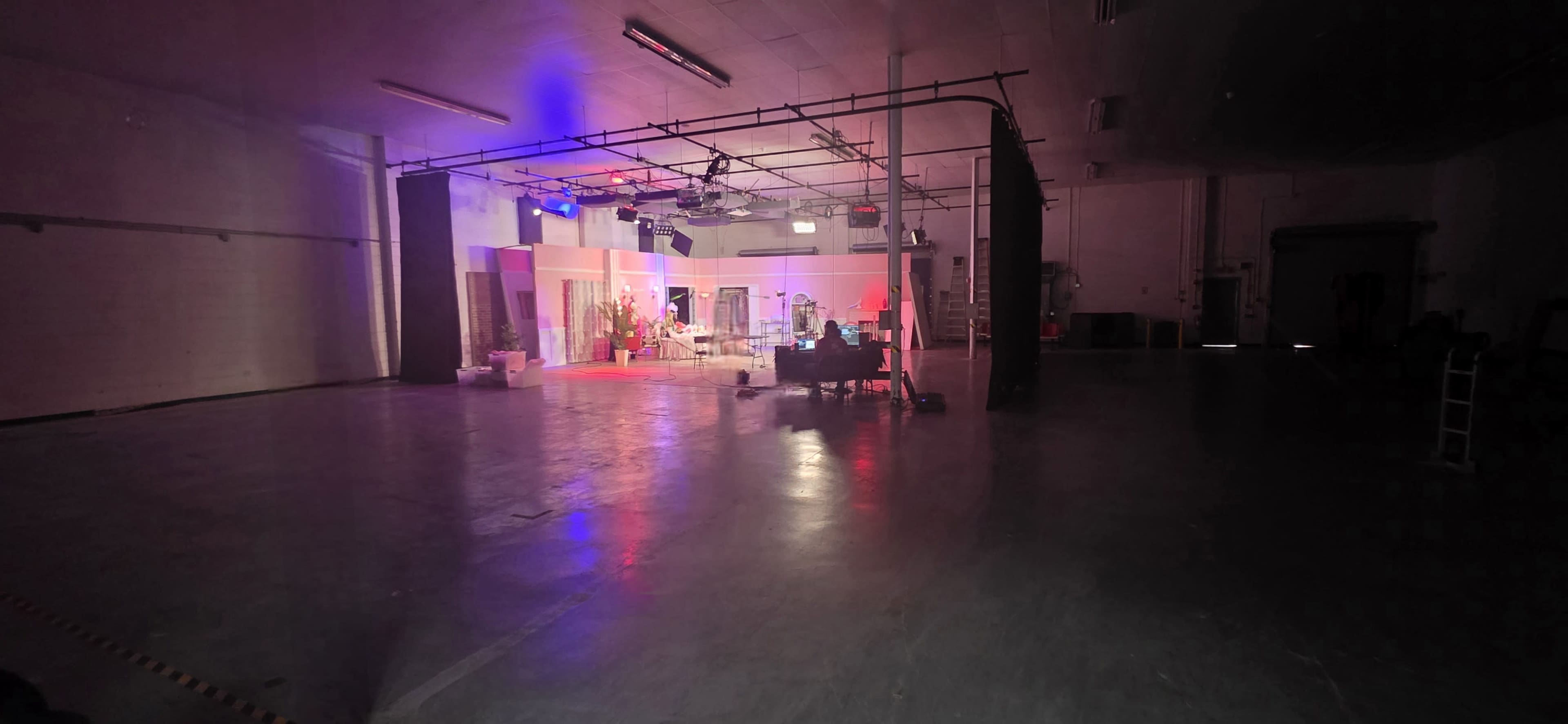 A large, empty studio space is illuminated with colorful lights, featuring a set arrangement and equipment in the background.