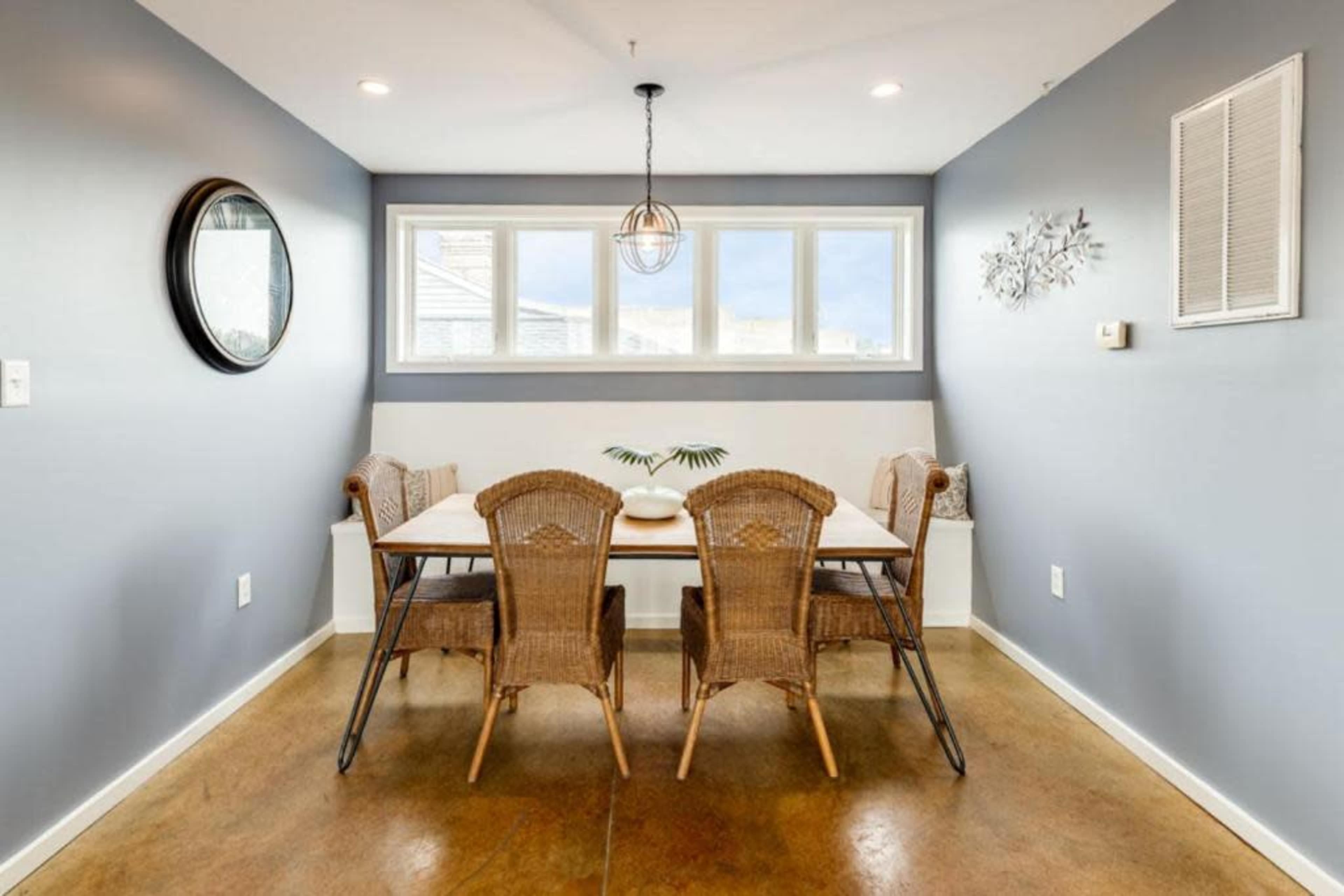 A dining area features a rectangular table surrounded by six wicker chairs, with a large window illuminating the space and gray walls providing a backdrop.