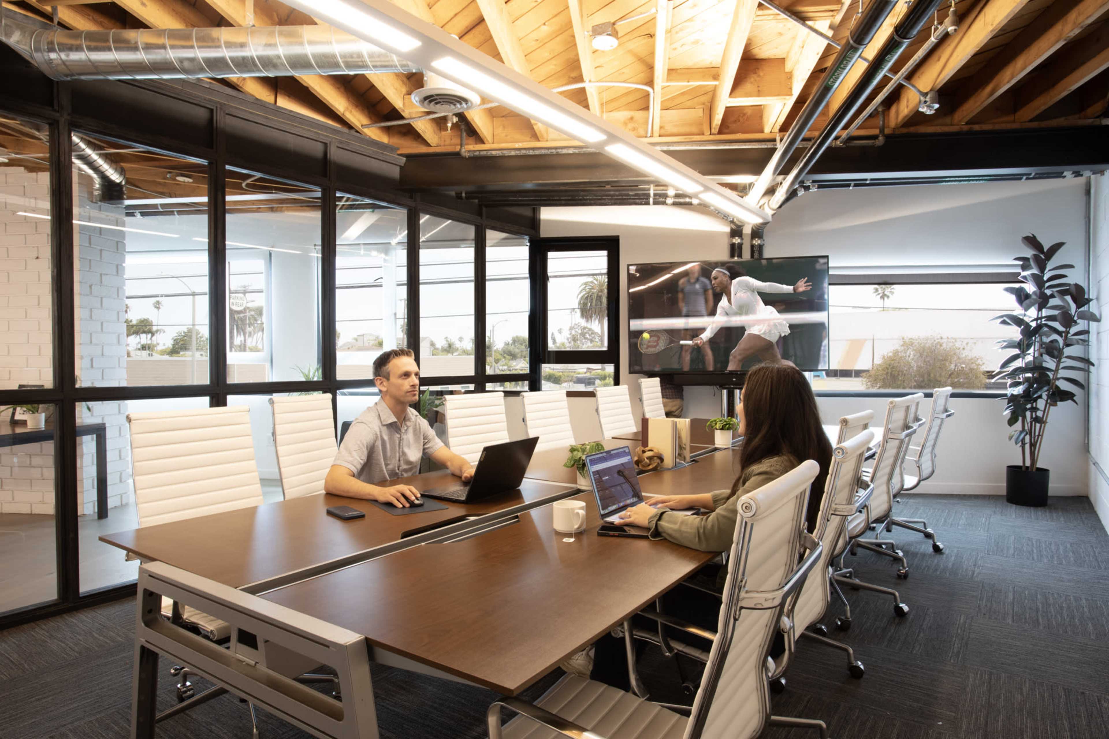 A modern conference room features a long wooden table, a laptop-wielding man, a woman using a laptop, and a large screen displaying a presentation.