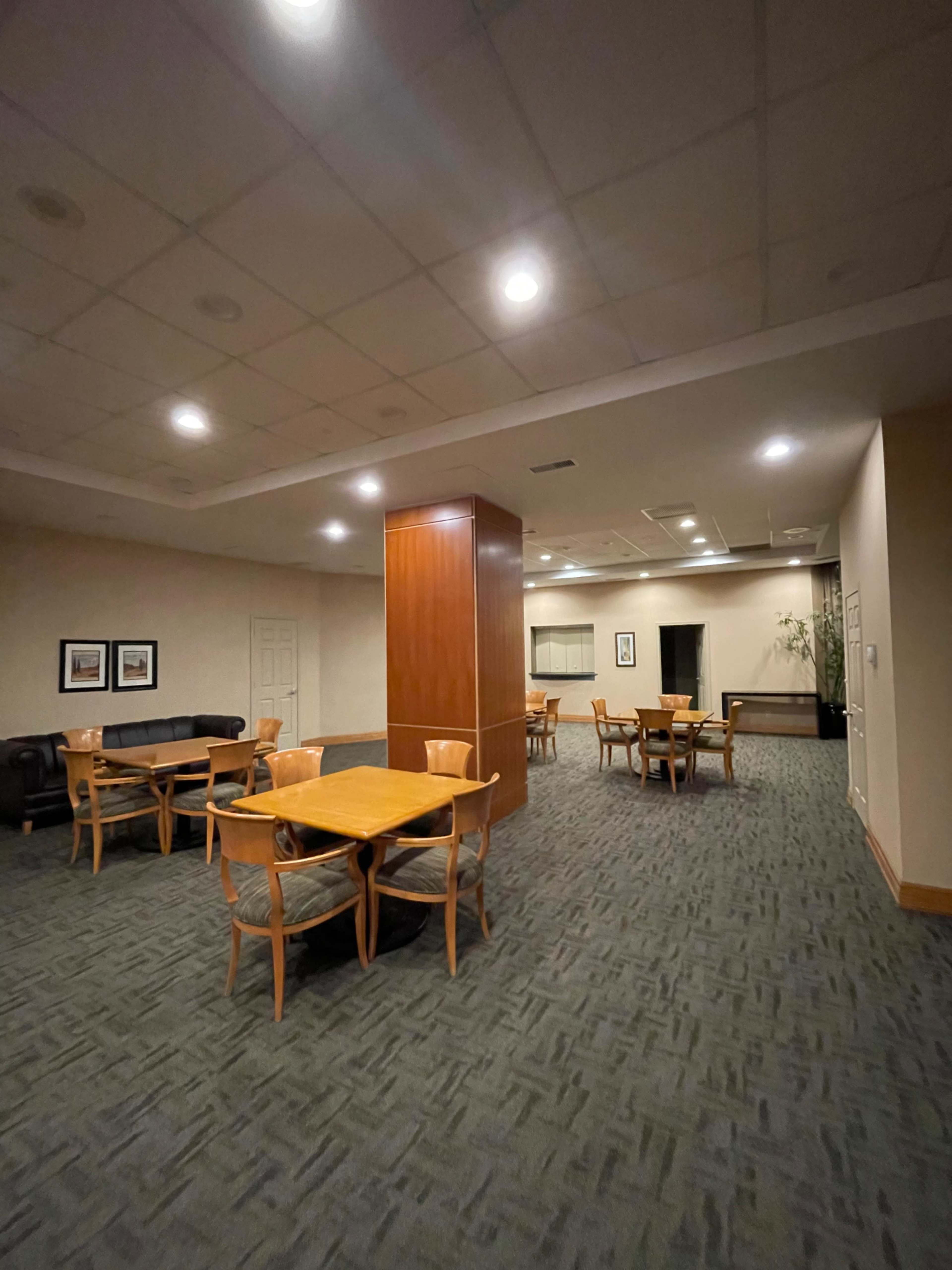 The image shows a spacious room with several wooden tables and chairs arranged around a central column, with a couch and a plant in the background.