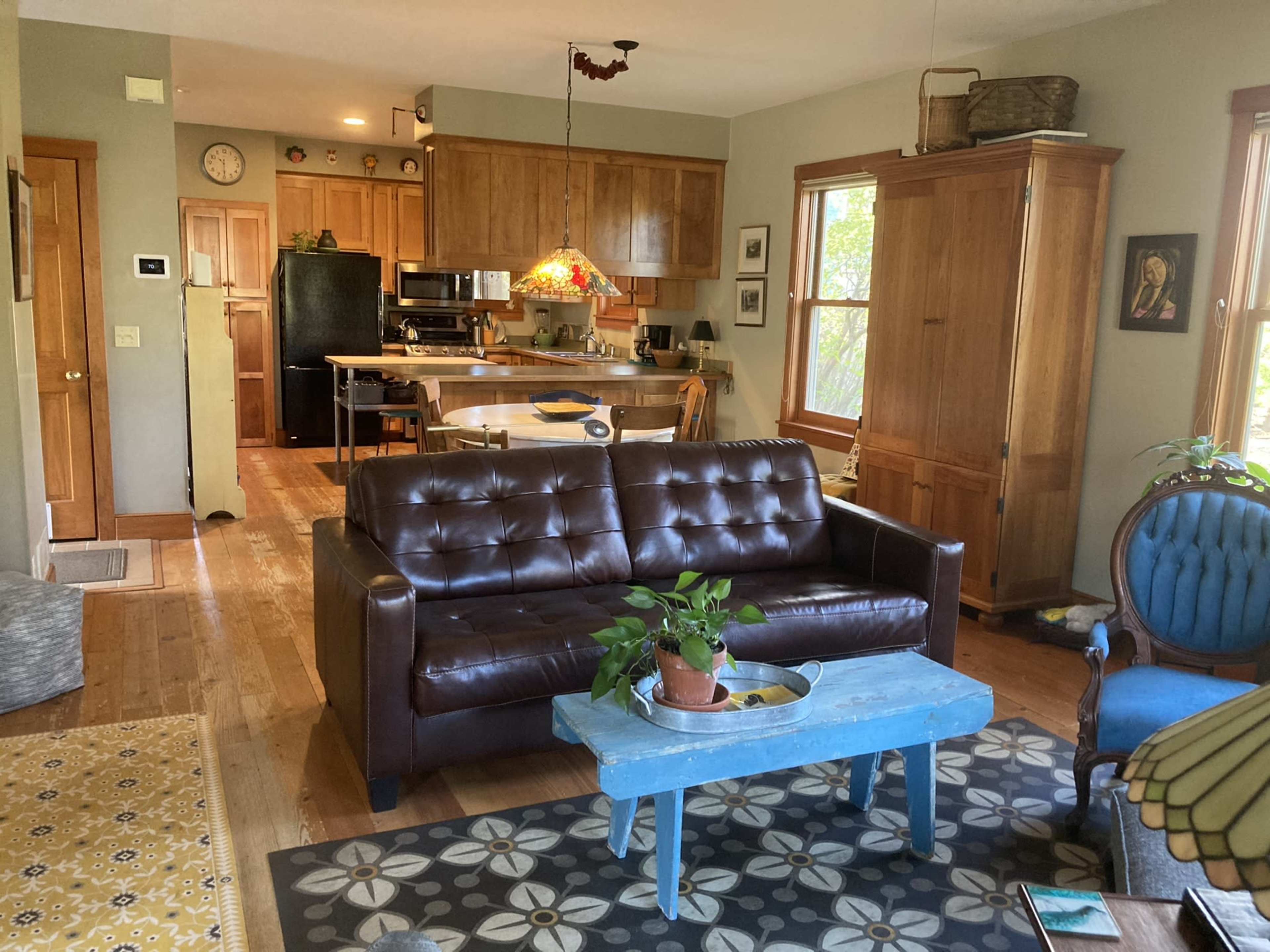 A cozy living room connected to a kitchen, featuring a brown leather sofa, a blue coffee table with a plant, and wooden cabinetry in the background.