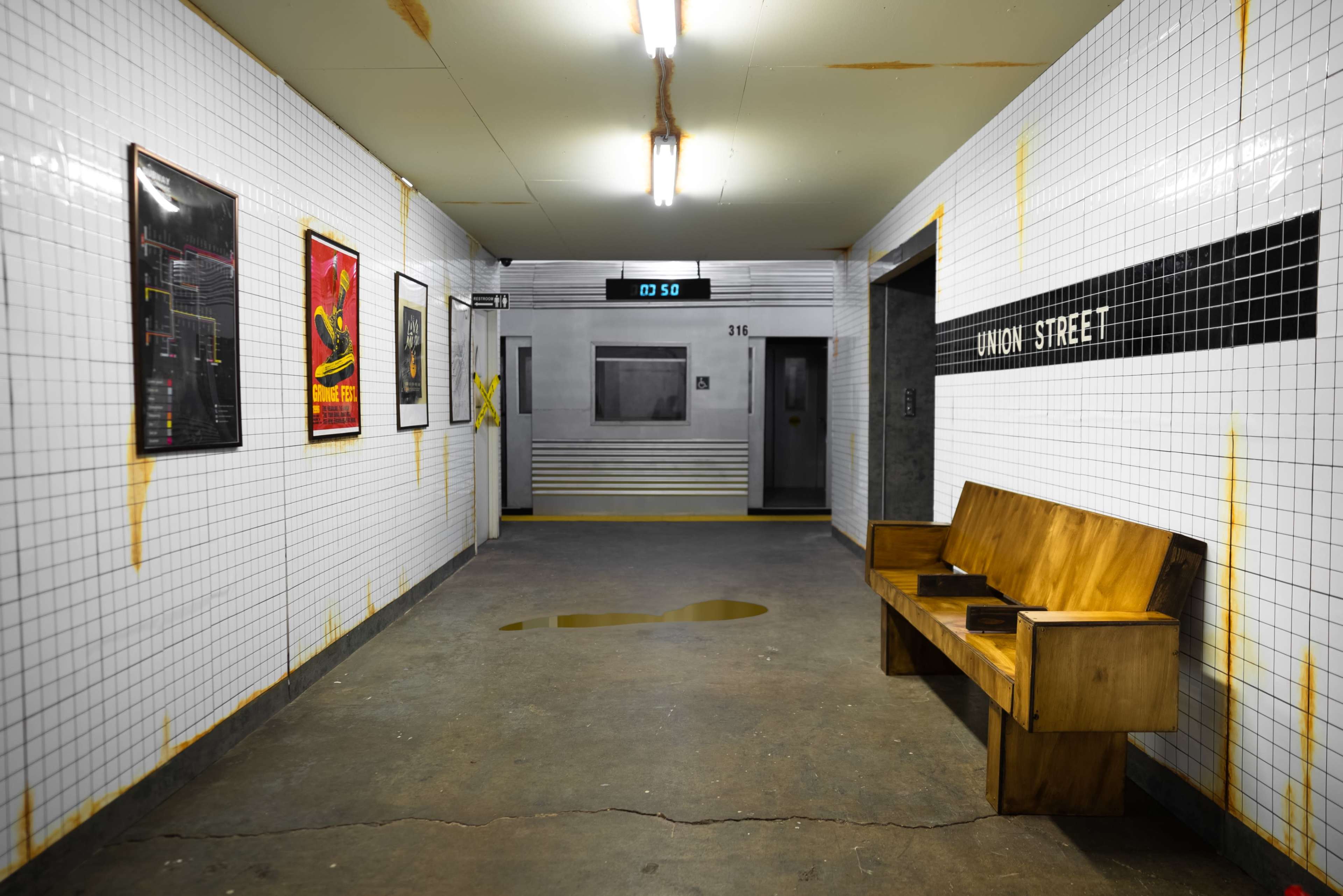 A tiled corridor in a subway station with a wooden bench, elevator doors, and a digital display indicating the time.