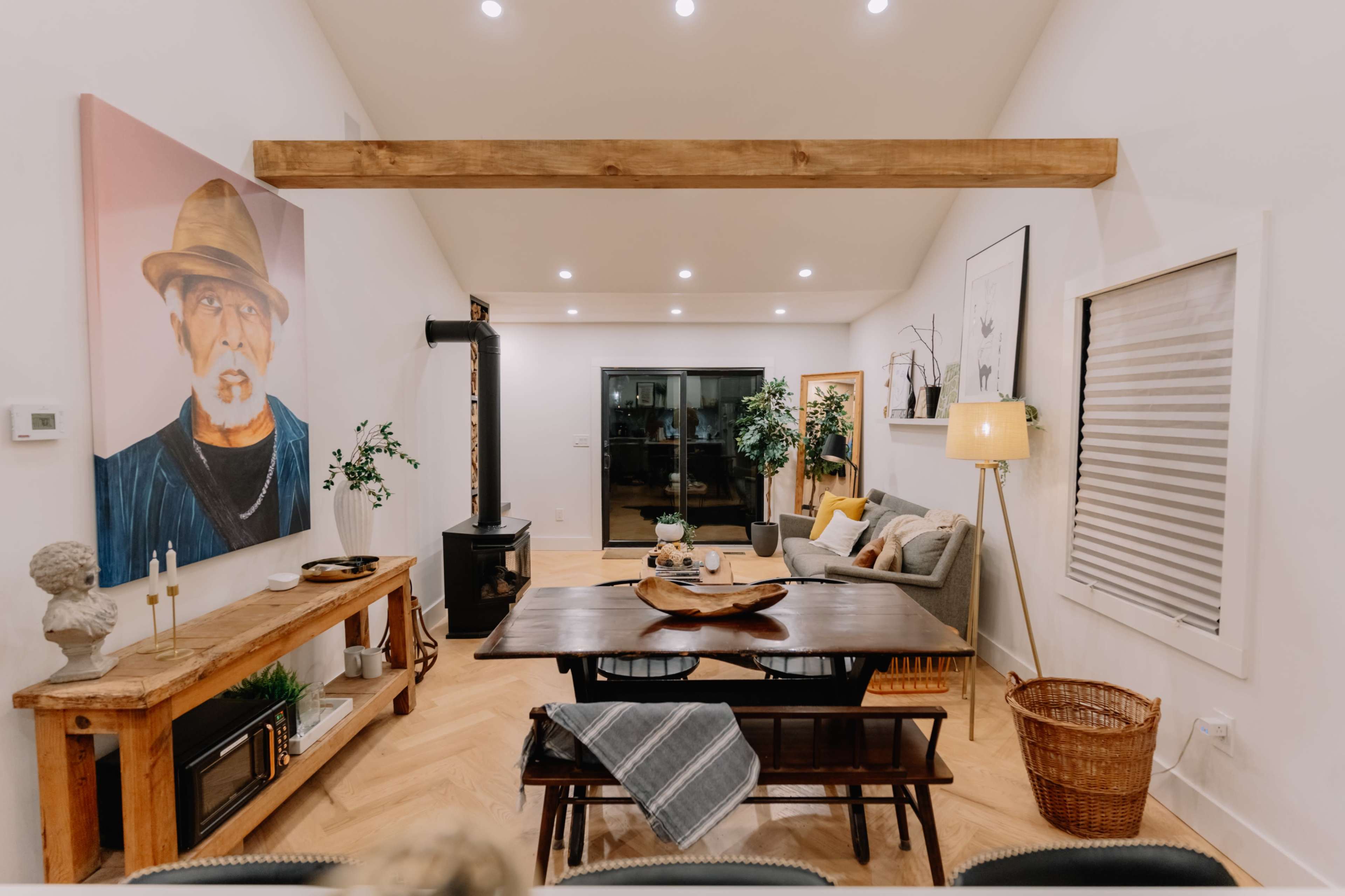 The image shows a modern living room with a dining area, featuring a large artwork on the wall, a wooden table, a black stove, and various plants and decorative items.