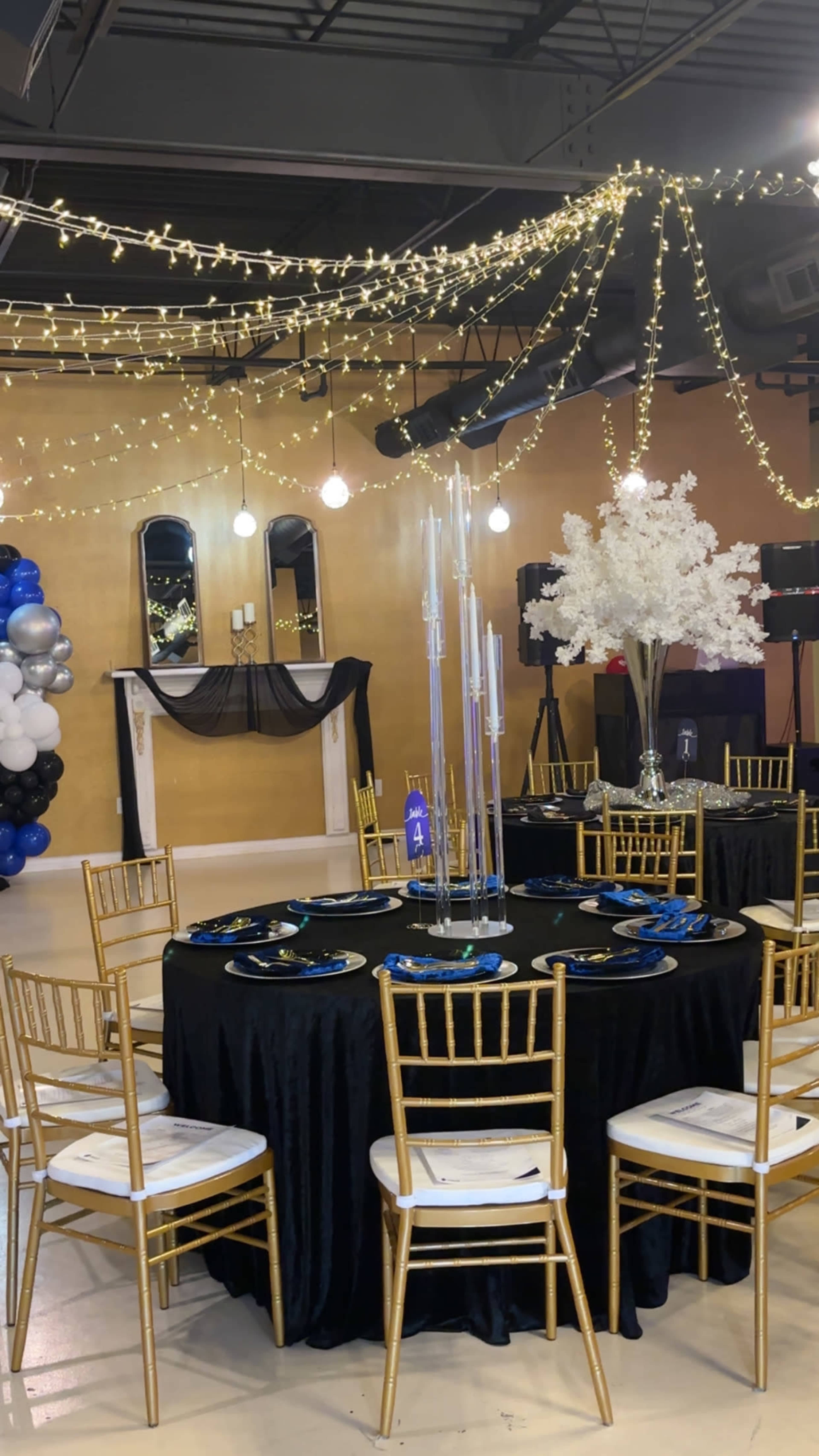 The image shows a banquet hall decorated for an event, featuring round tables with black tablecloths, gold chairs, and elegant centerpieces, surrounded by string lights and a backdrop with balloons.