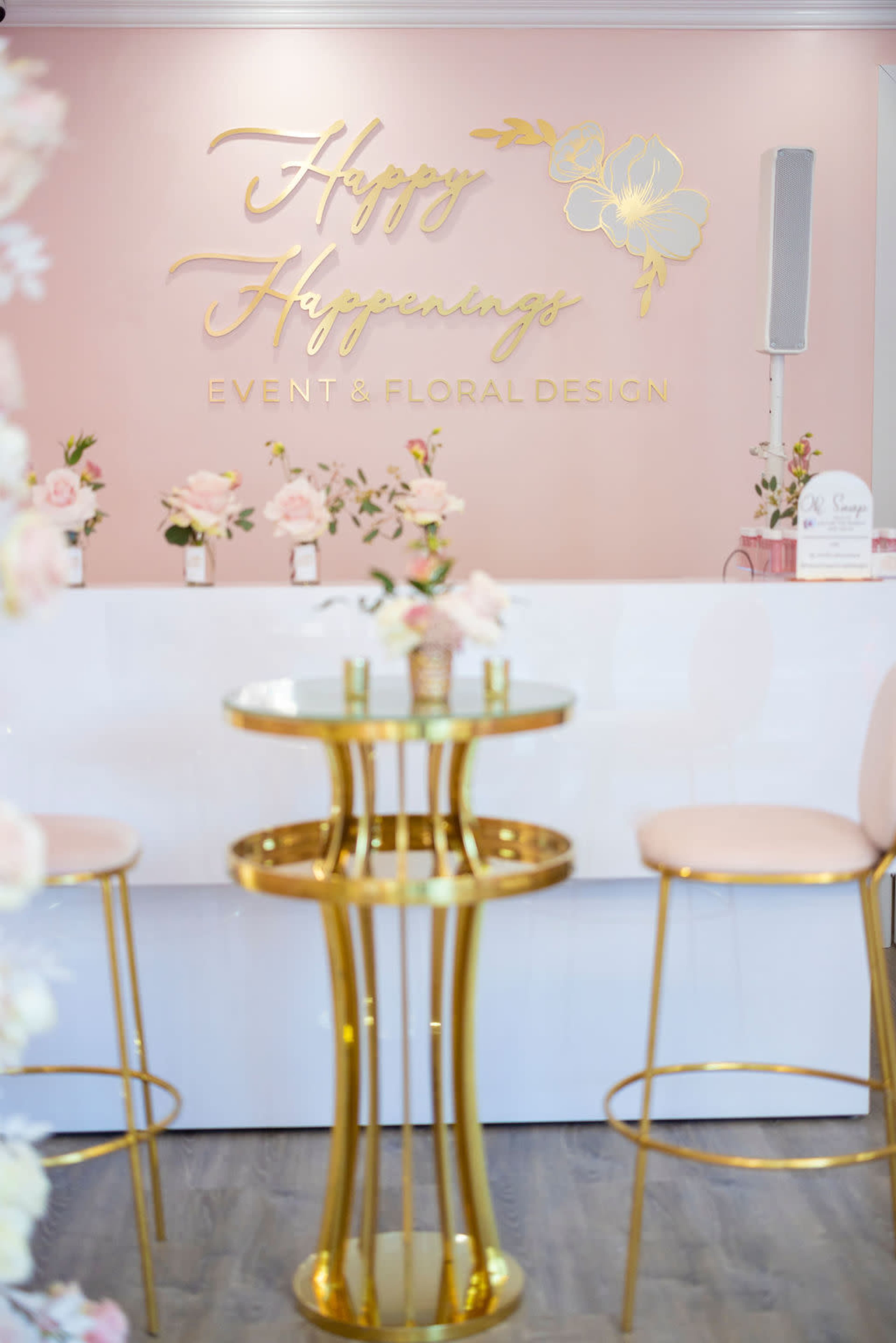 The image shows a pink wall with a gold sign that reads "Happy Happenings Event & Floral Design" above elegant tables adorned with floral arrangements.