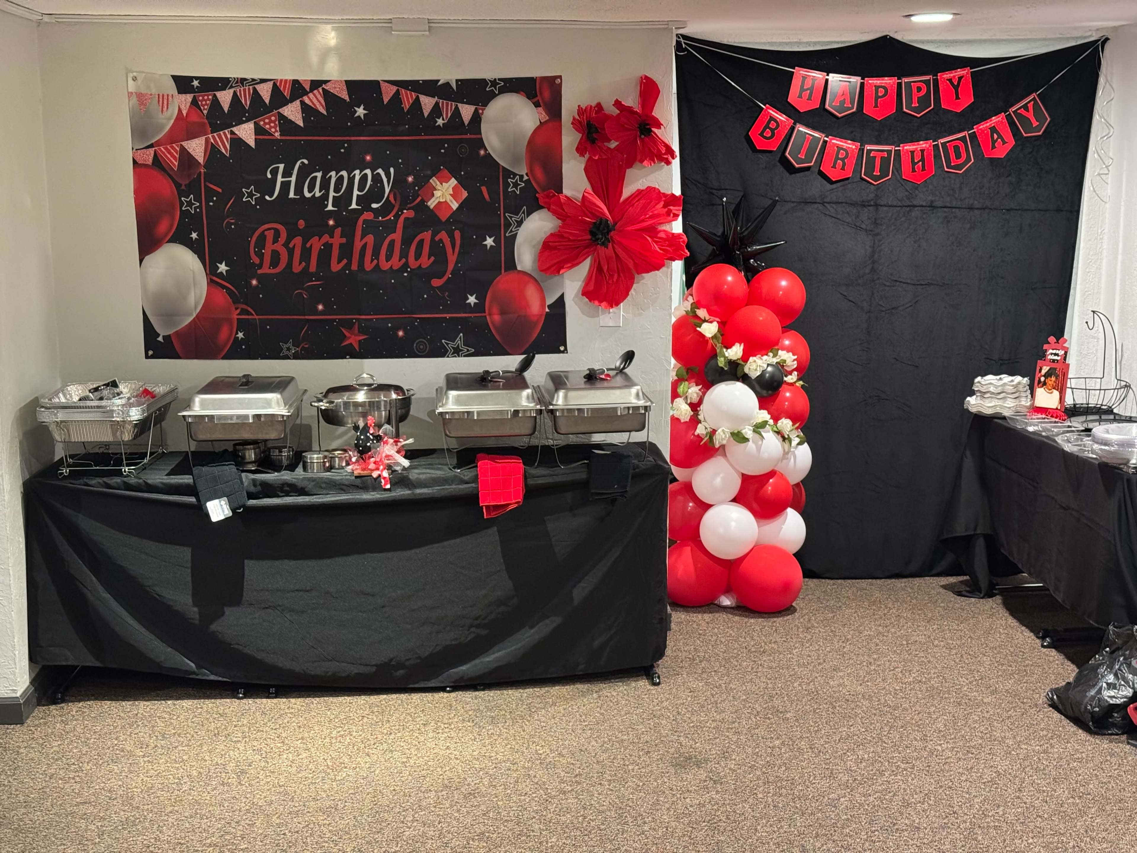 A birthday-themed buffet setup is decorated with balloons and banners, featuring food warmers and a black tablecloth on one side.