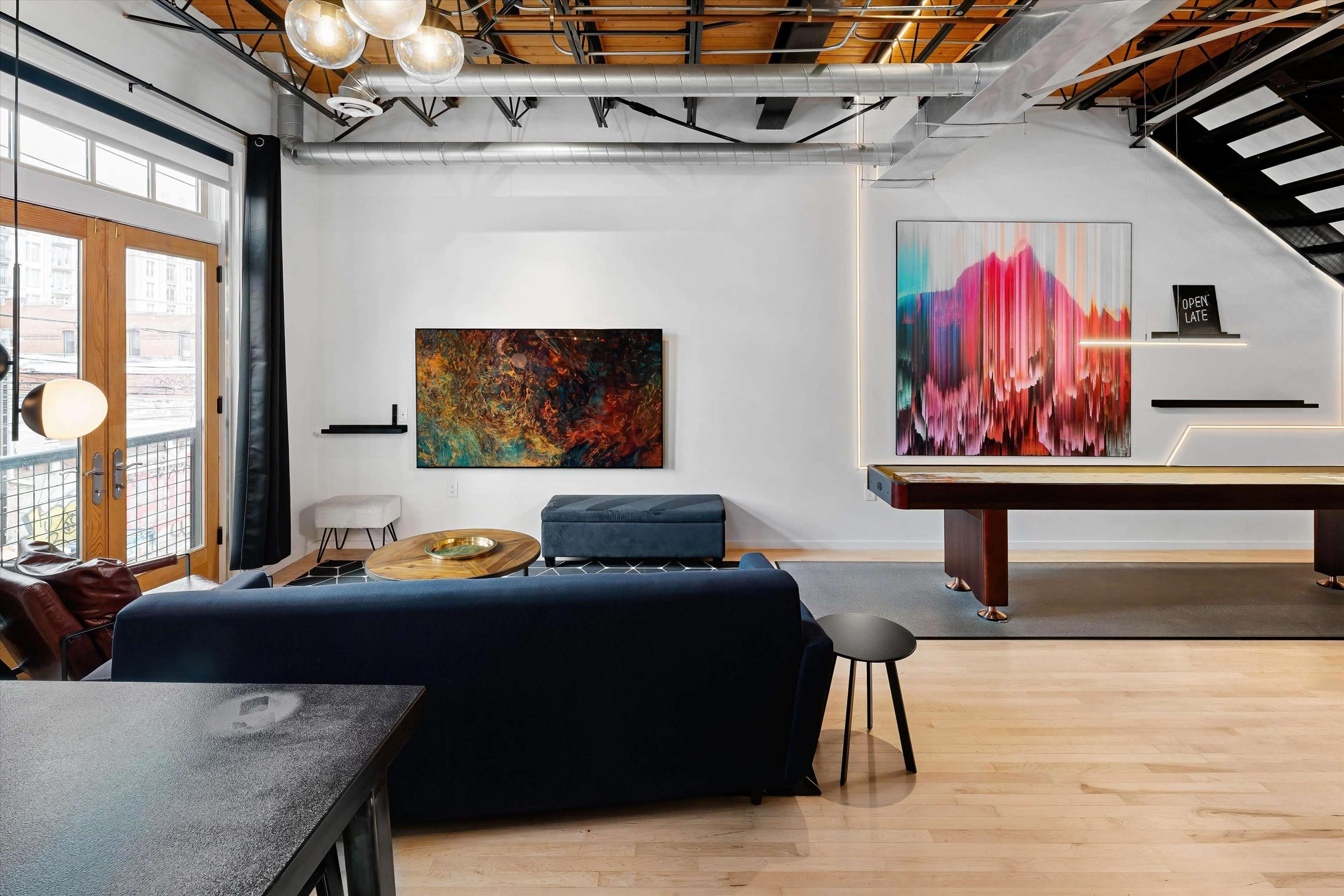 The image shows a modern living space featuring a blue sofa, a round wooden coffee table, colorful wall art, and a pool table, all under warm lighting with an open layout.