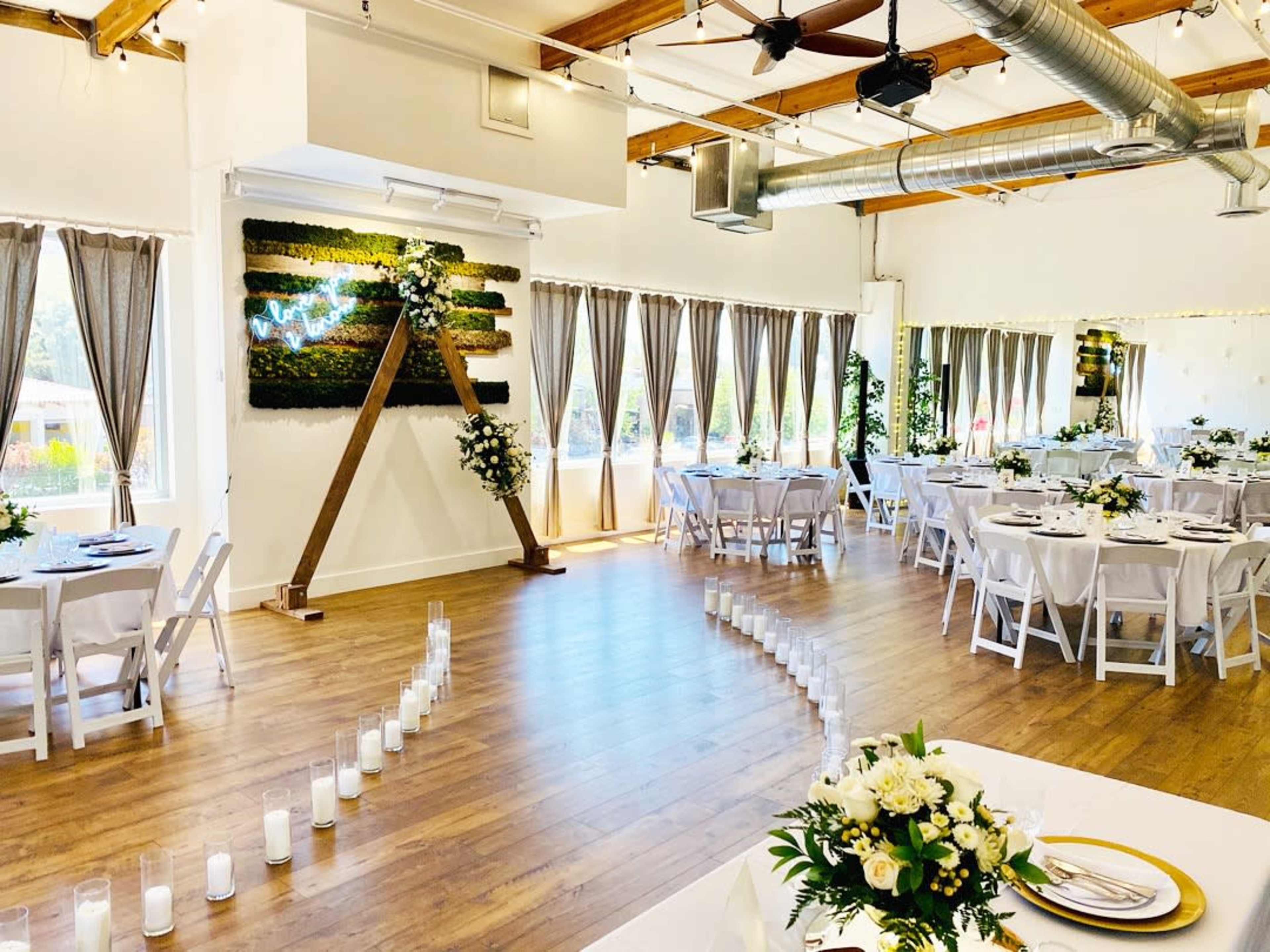An elegantly decorated event space with tables set for a gathering, featuring a floral arrangement and a walkway lined with candles.