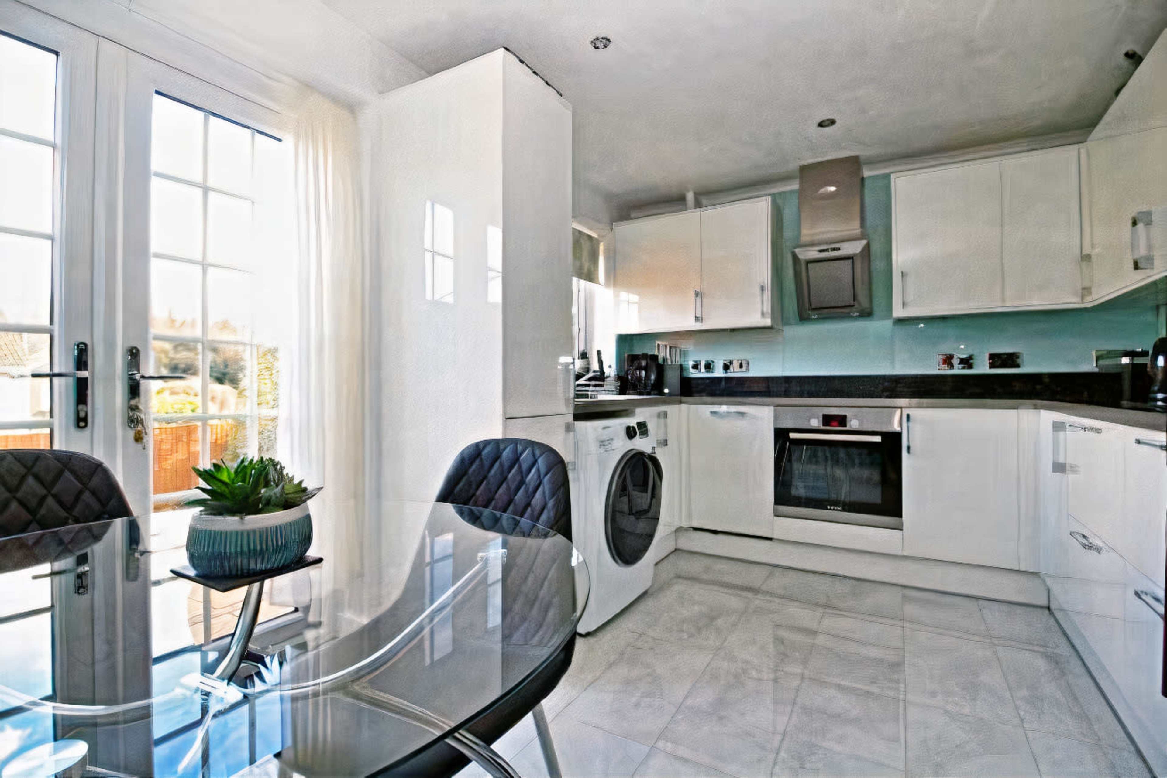 A modern kitchen features white cabinets, stainless steel appliances, a washing machine, and a glass dining table with a small plant.