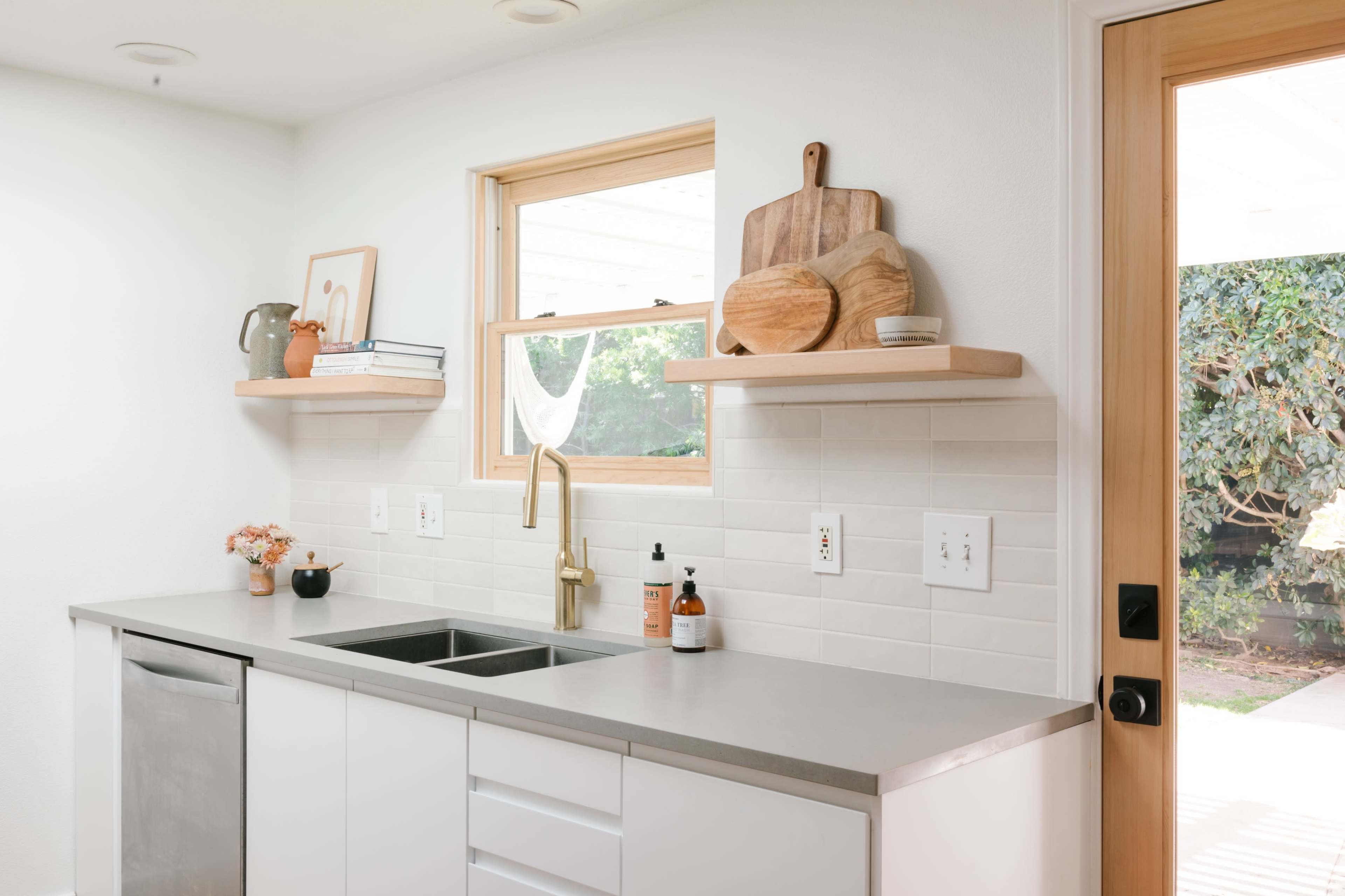 The image shows a modern kitchen featuring a stainless steel sink, a minimalist countertop, and open shelves displaying wooden cutting boards and decorative items.