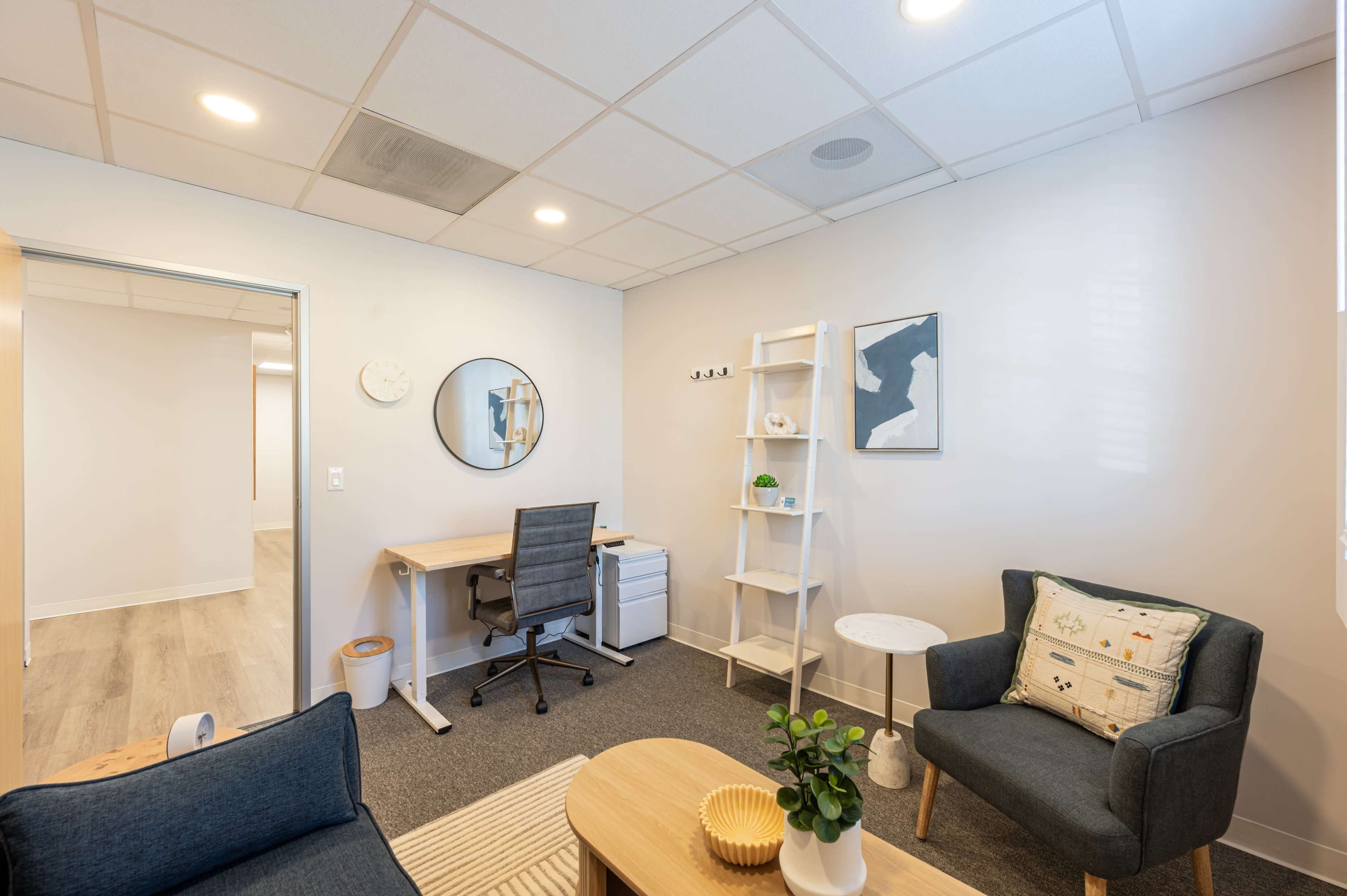 The image shows a modern office space with a desk, chair, shelving unit, and seating area featuring a small table and decorative items.