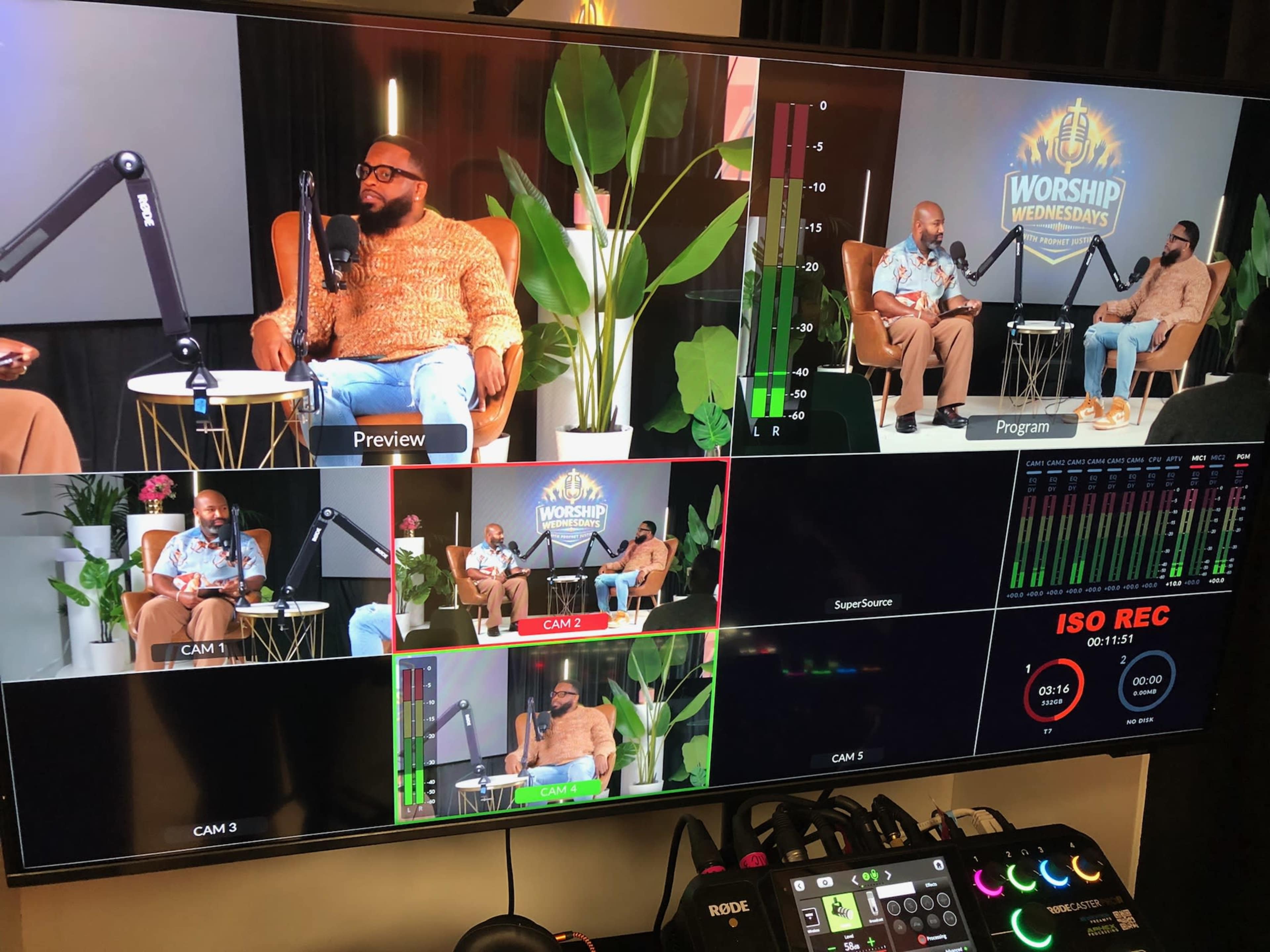 The image shows a television studio setup with multiple screens displaying a recording of a discussion, featuring two individuals seated on chairs surrounded by indoor plants.