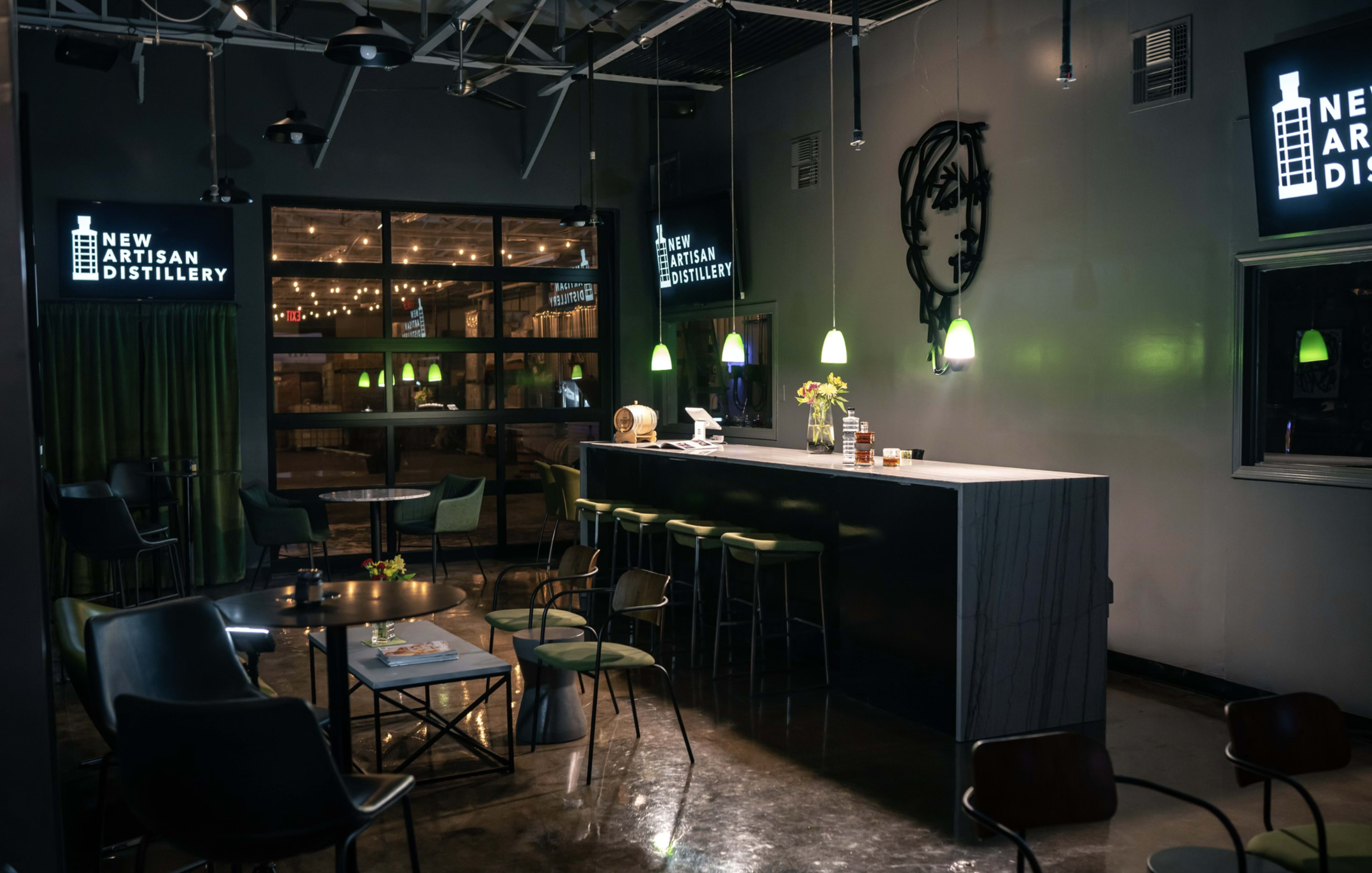A dimly lit distillery interior featuring a long bar with modern stools and tables under illuminated signs.