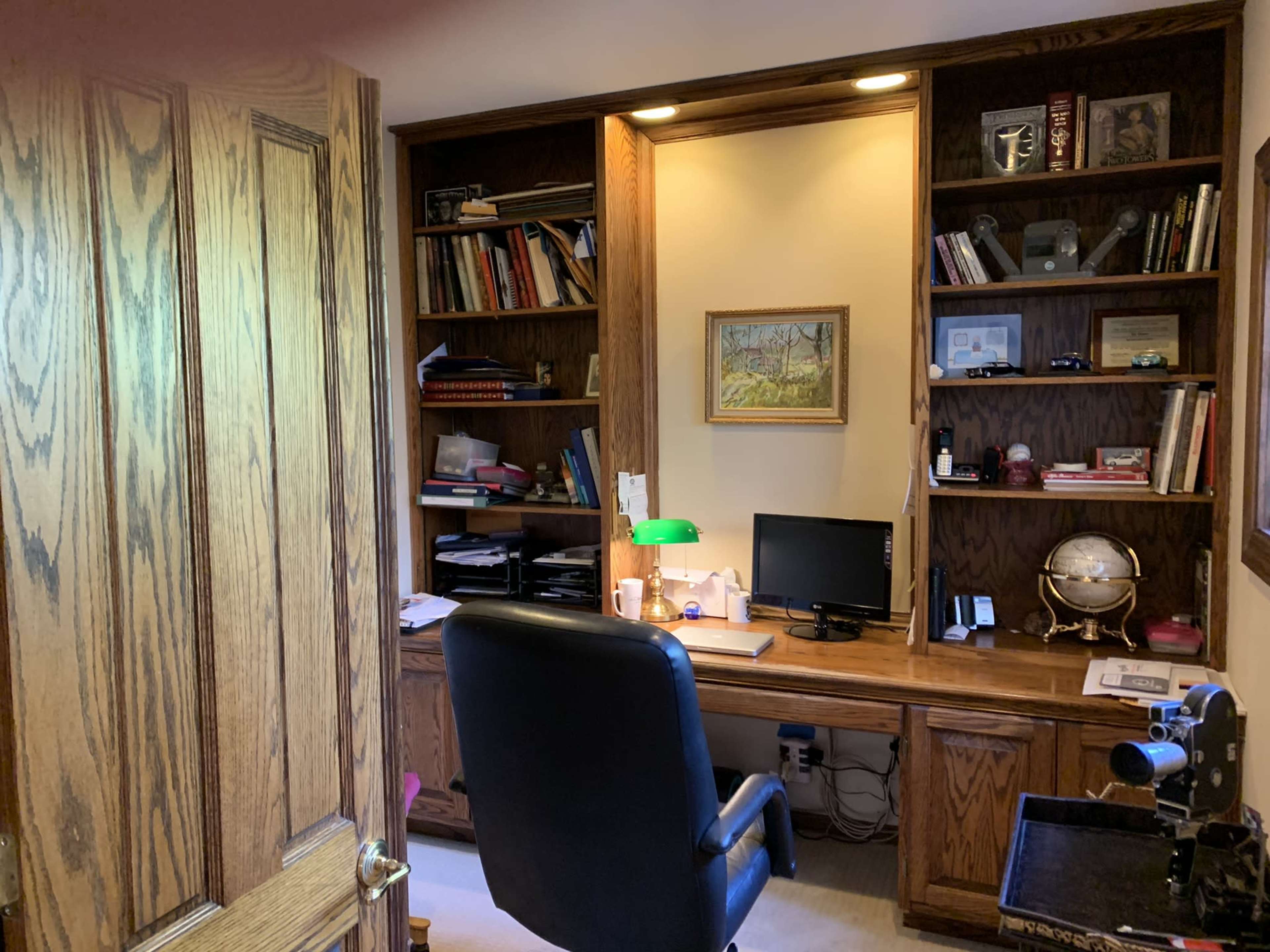 A home office with wooden built-in shelves filled with books, a desk with a computer and a green lamp, and a black chair facing the workspace.
